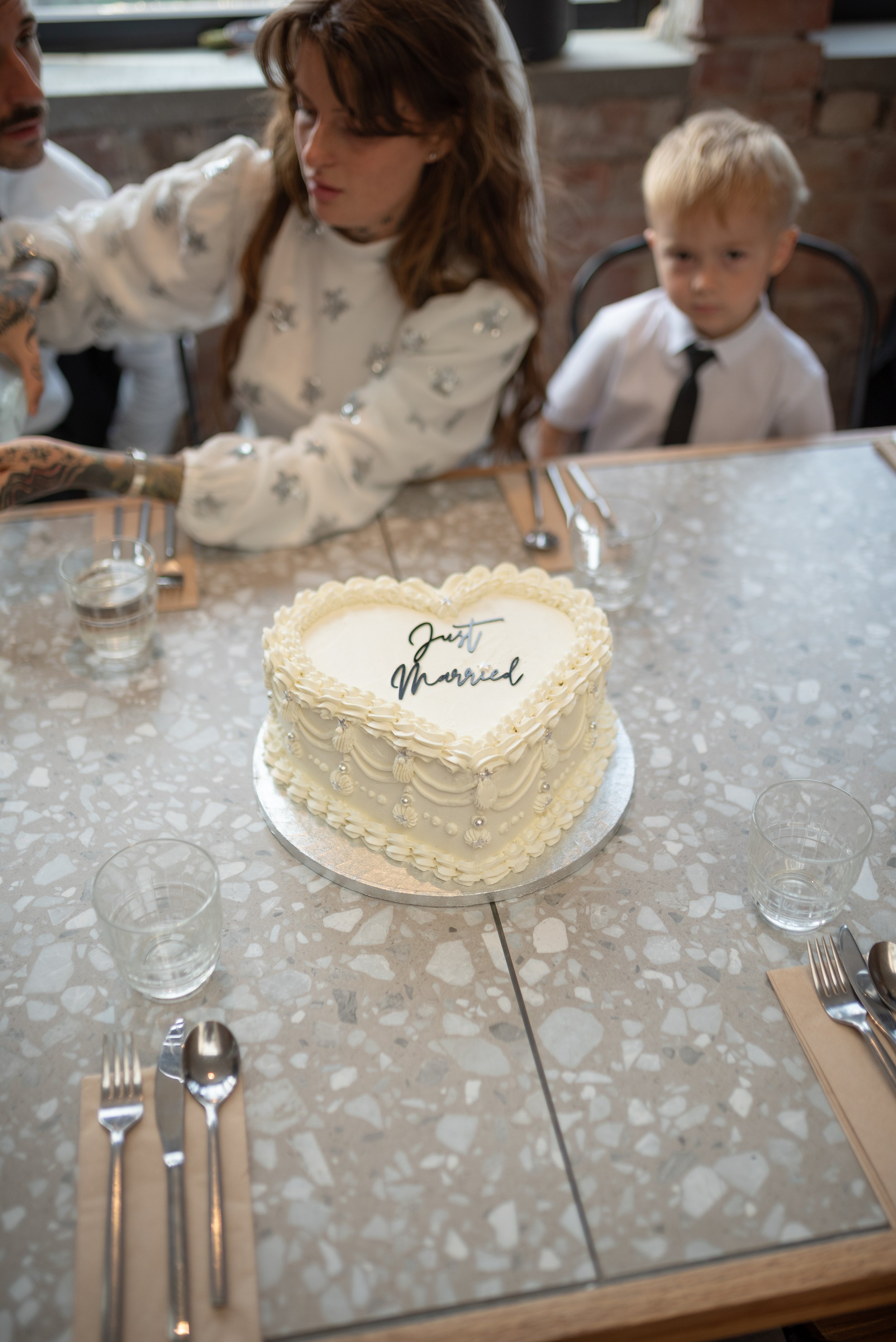 A heart-shaped wedding cake with white icing and decorative piping, featuring the words 'Just Married' on top, sits on a table surrounded by empty glasses and cutlery. In the background, a woman with long brown hair and a white dress, and a young boy