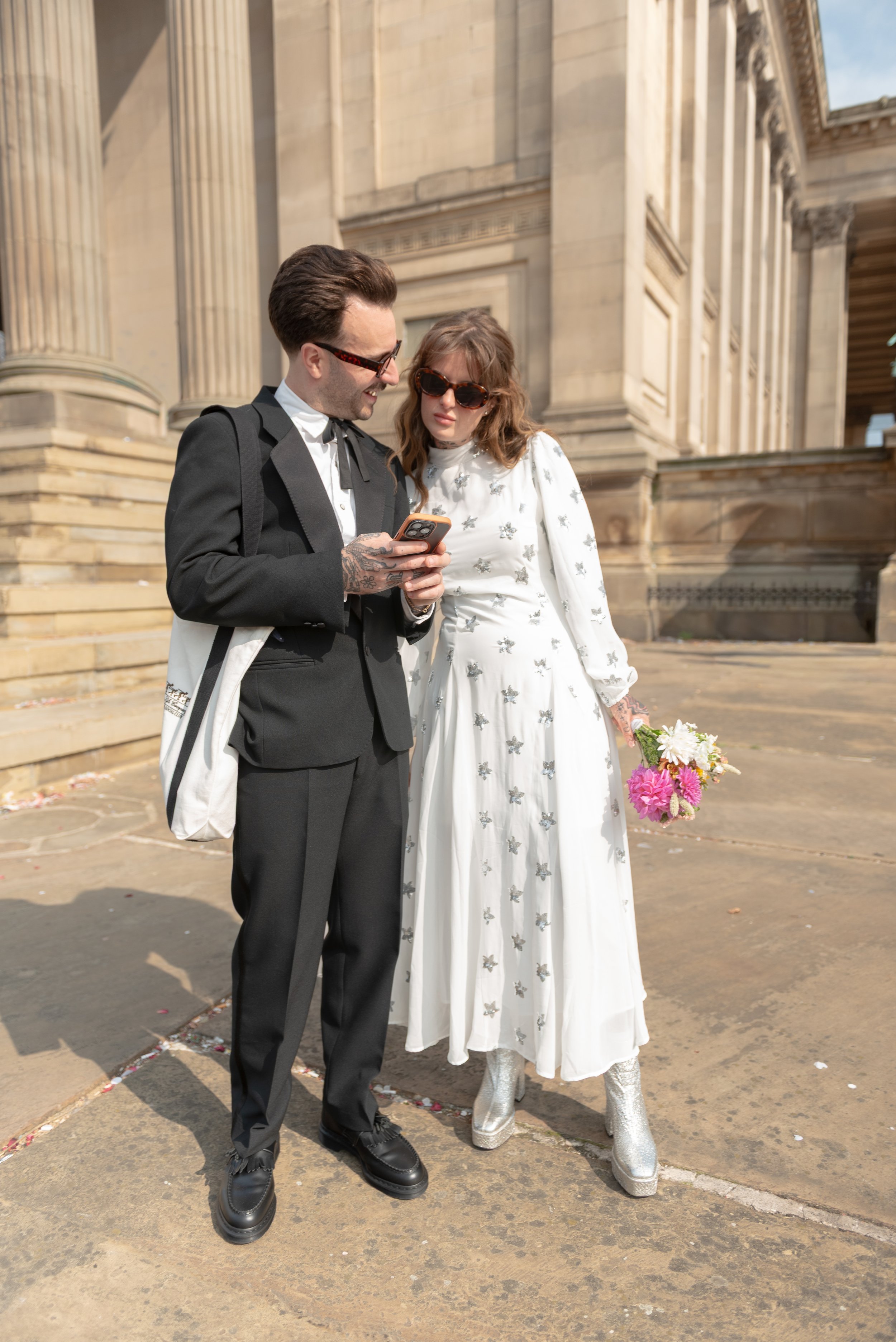 A man and woman dressed in formal attire standing outside a historic stone building, looking at a smartphone together, with the woman holding a bouquet of flowers and wearing silver platform boots.