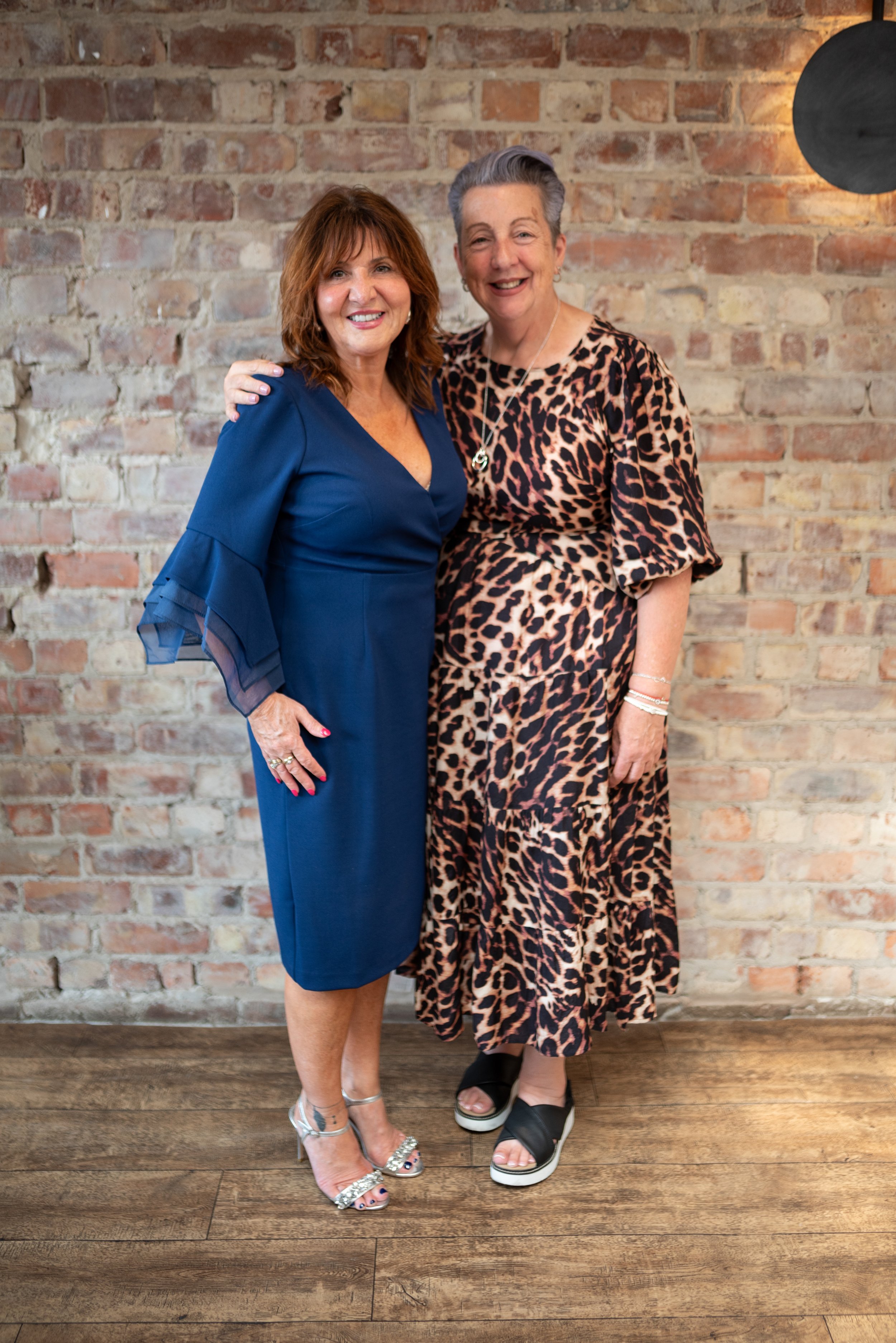 Two women smiling and posing together in front of a brick wall, one in a blue dress and the other in a leopard print dress.