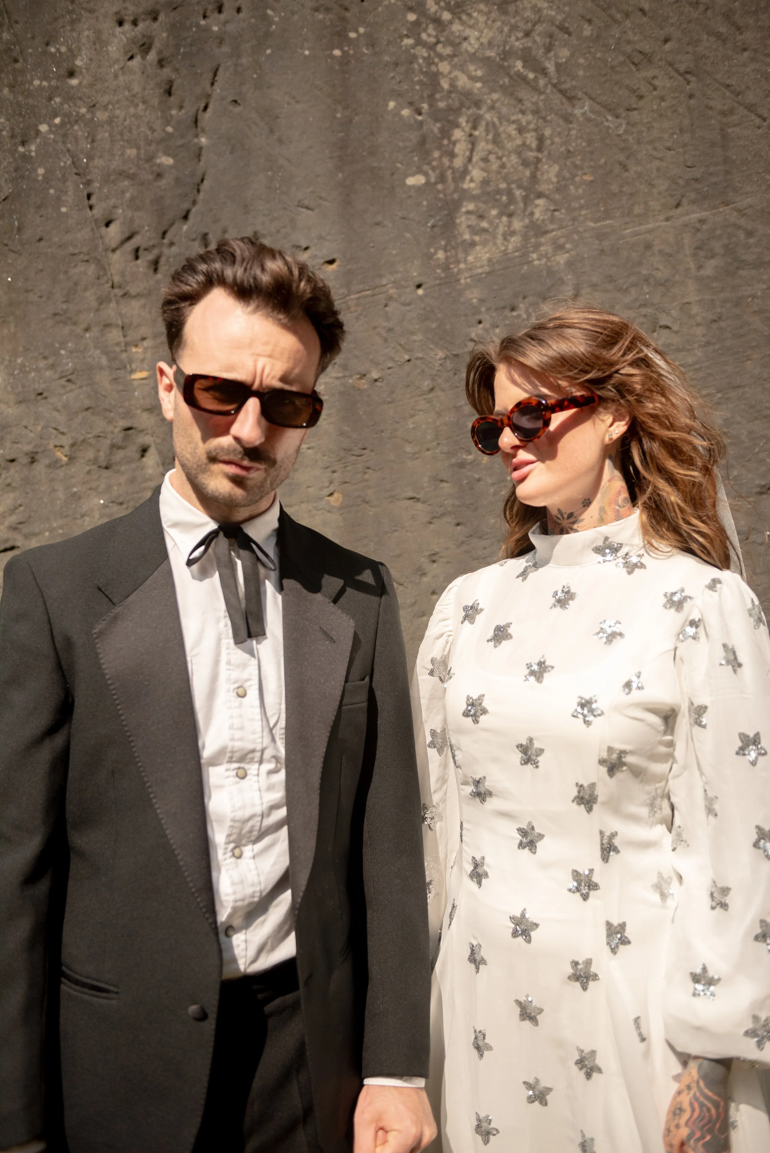 A man and woman standing in front of a stone wall, both wearing sunglasses. The man is dressed in a black suit with a white shirt and a bow tie, with short, dressed hair. The woman is wearing a white dress with silver butterfly embellishments, large 