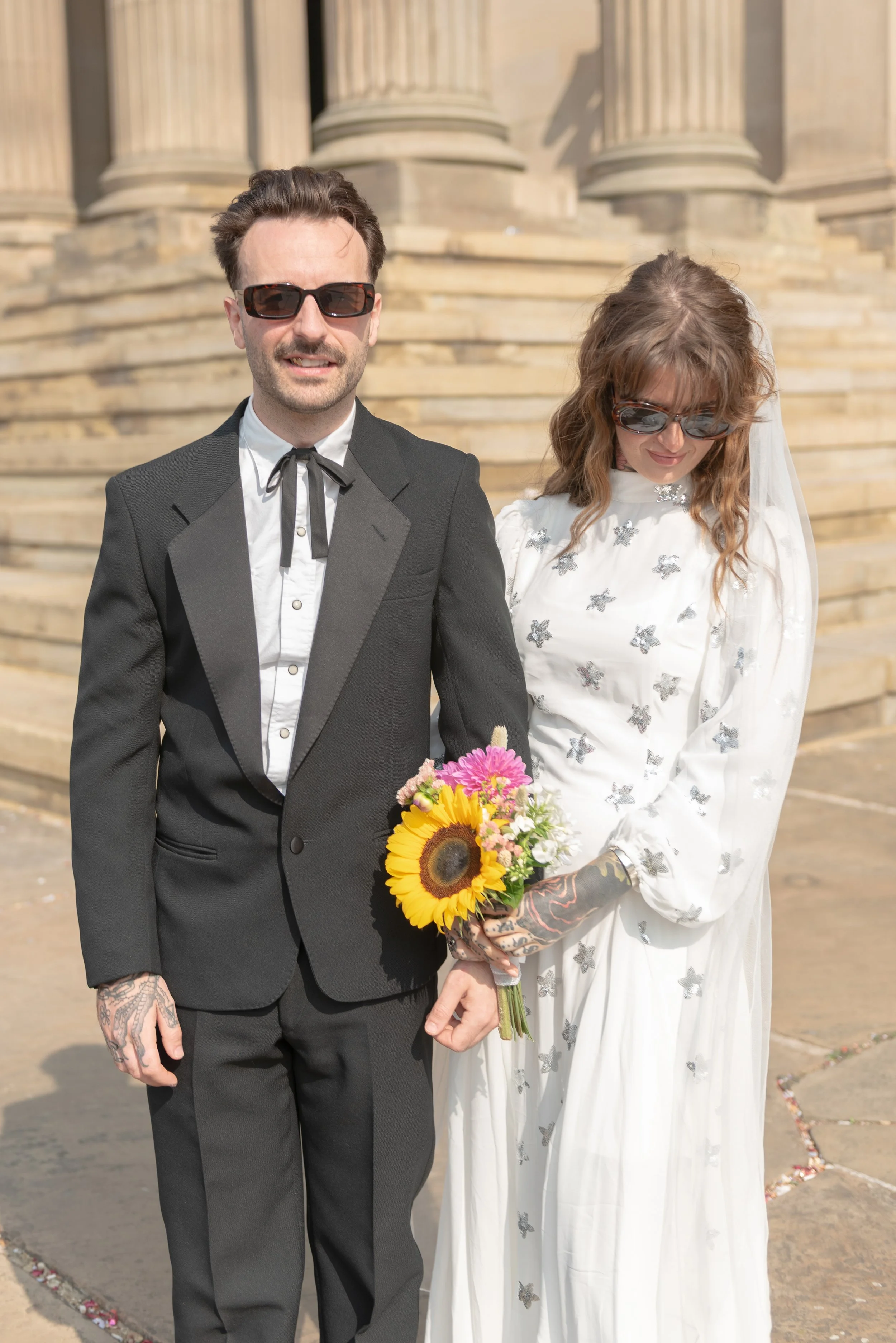 A couple dressed as a groom and bride, with the groom in a tuxedo and the bride in a white dress with flowers, standing outside in front of stone steps, both wearing sunglasses, holding a bouquet of flowers, with the bride looking down.