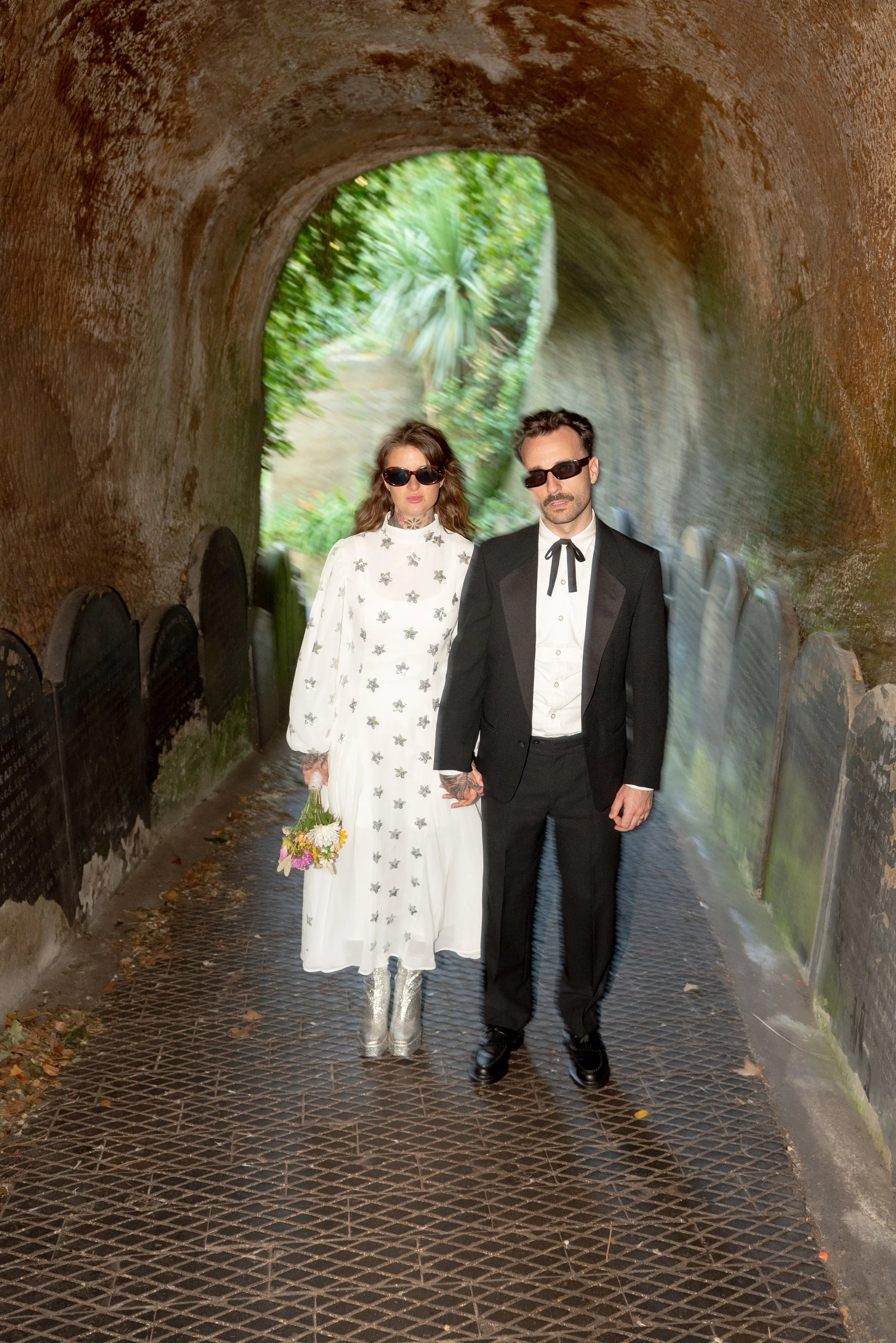 A woman and a man dressed in vintage-style clothing standing hand in hand in a tunnel with greenery visible at the end.