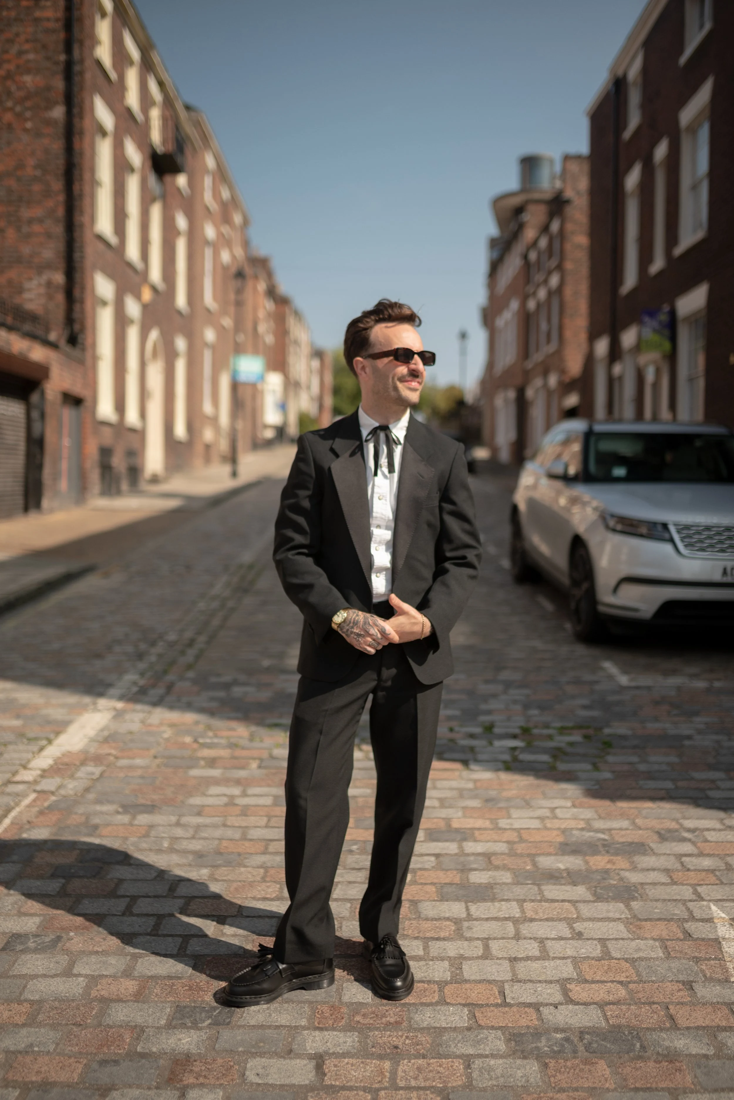 A man dressed in a black suit with a white shirt and black tie, wearing sunglasses, standing on a cobblestone street. He has tattoos on his hands and is looking to the side with a smile.