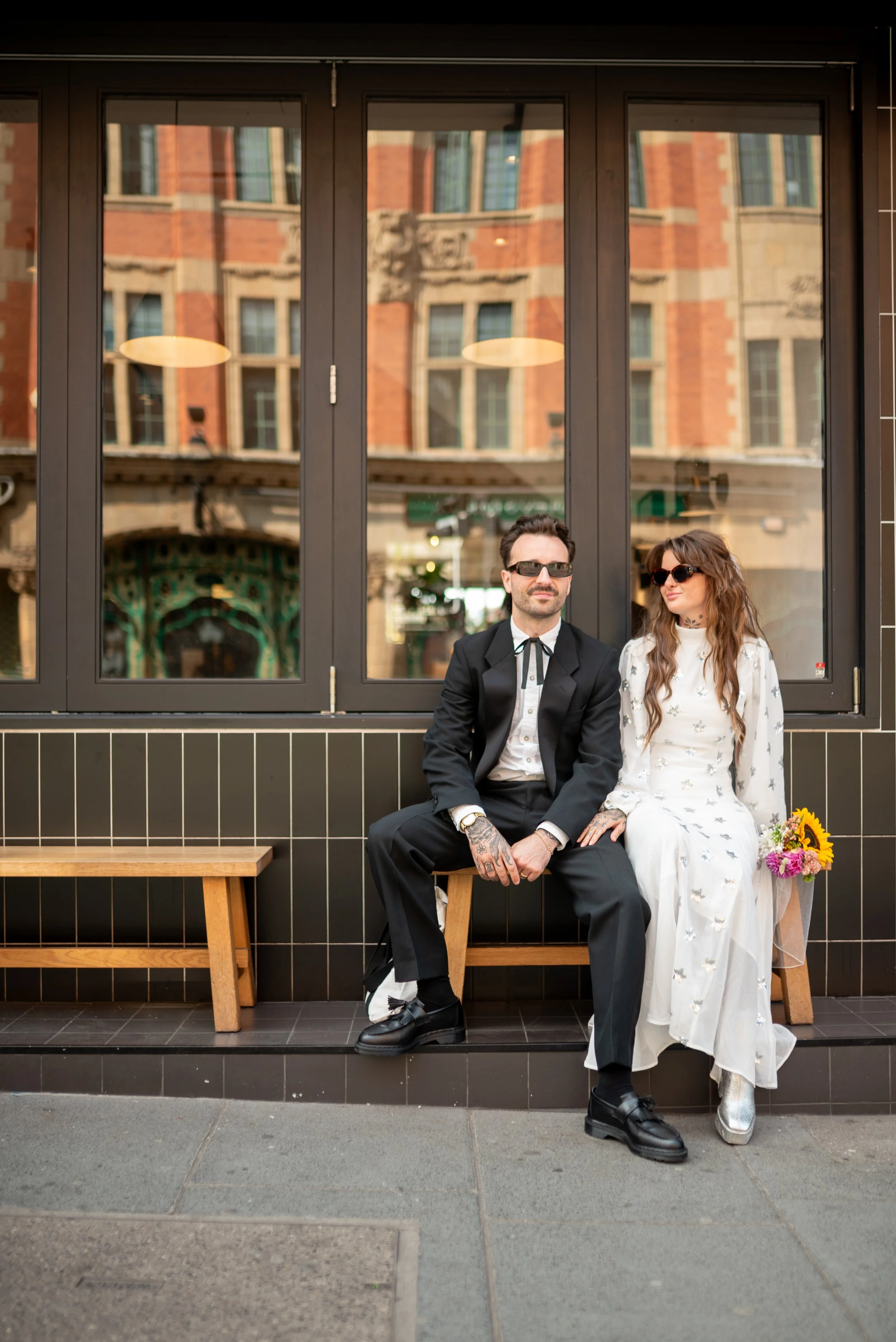 A man and woman sit on a wooden bench outside a building with large windows and a brick facade, wearing fashionable clothing and sunglasses, with a bouquet of flowers beside the woman.