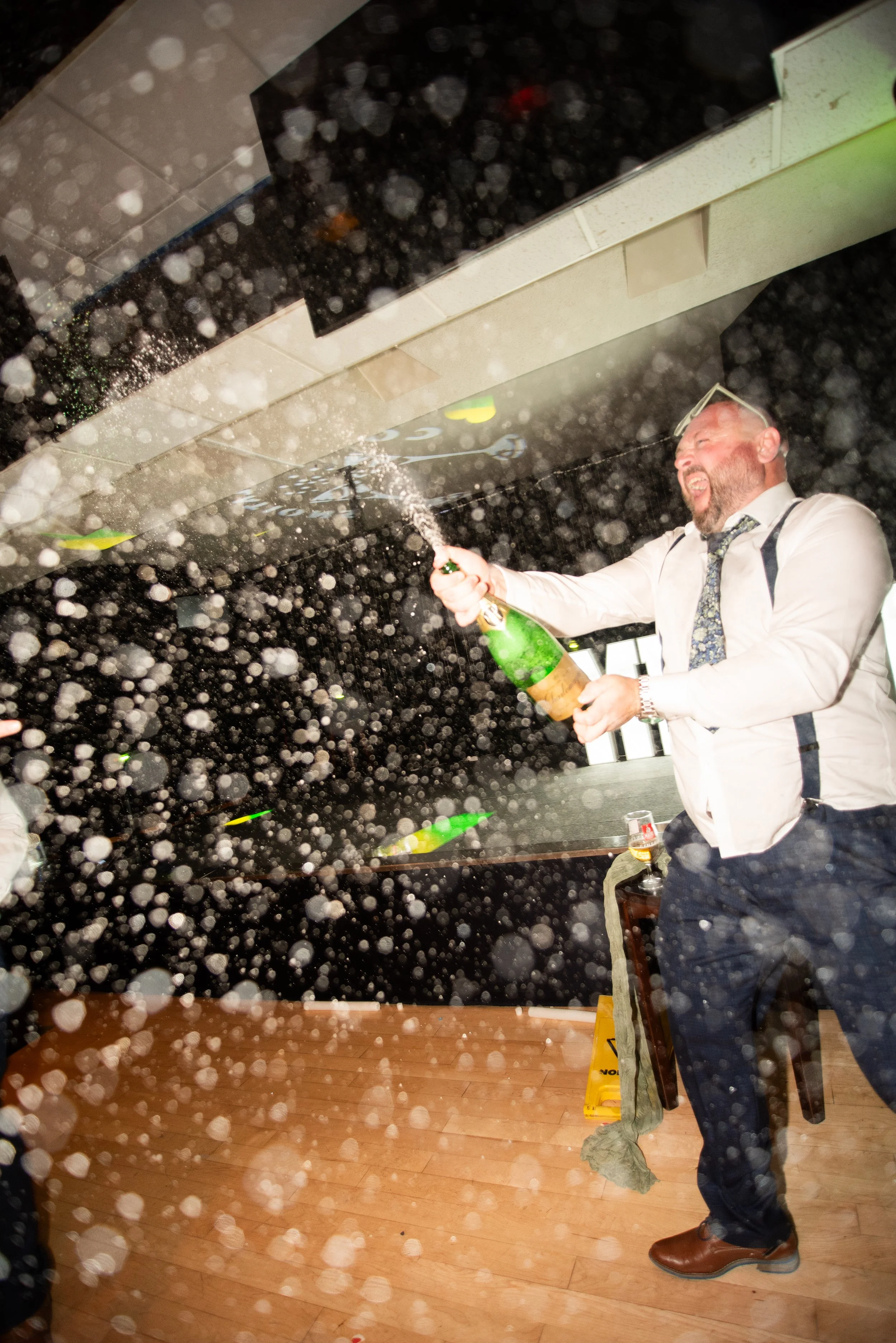 A man in a white shirt and dark pants celebrates with a bottle of champagne in a spray, with foam and bubbles all around, in a party setting.