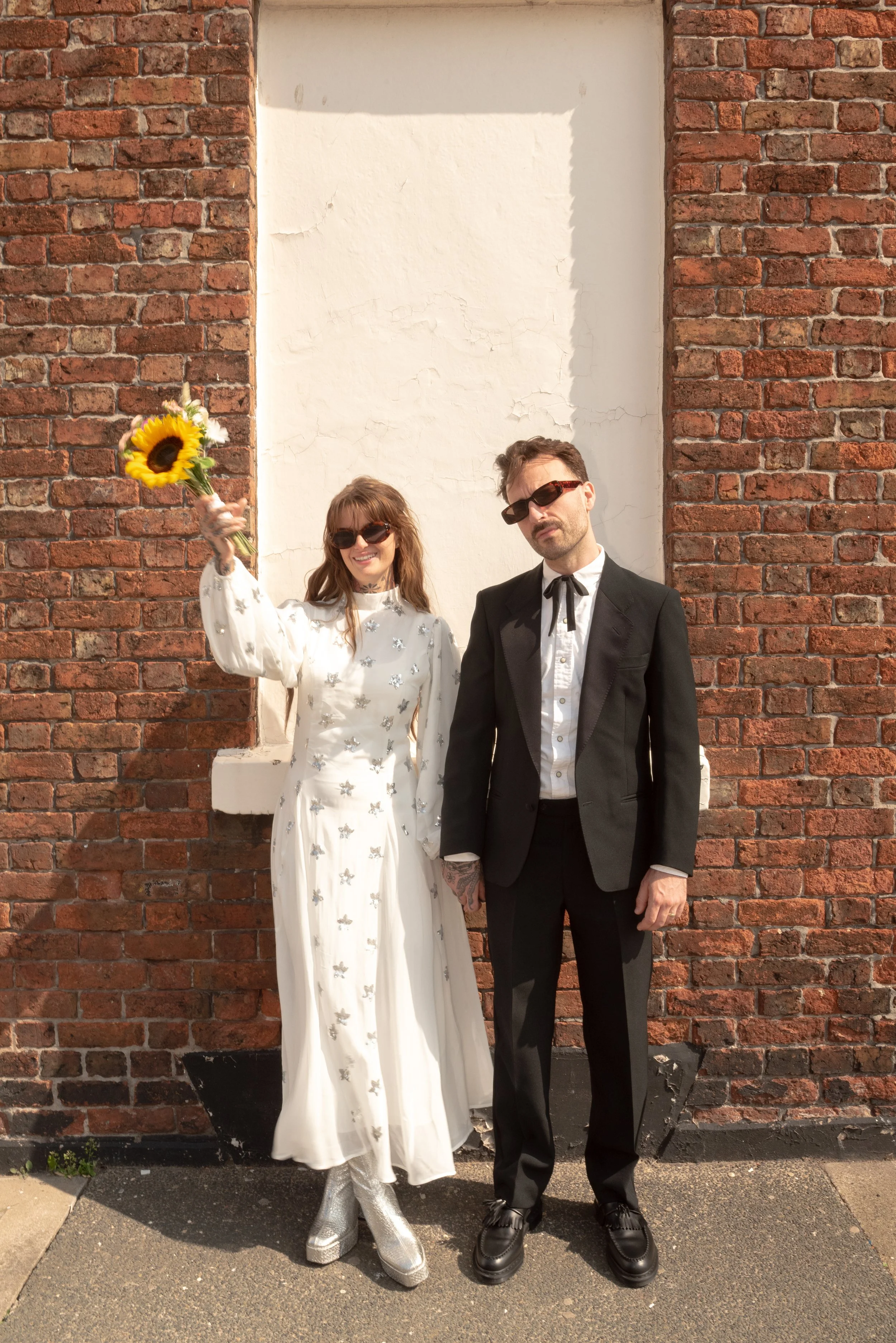 A woman and a man dressed in vintage-style clothing standing in front of a brick wall. The woman is holding a bouquet of sunflowers and smiling, while the man is wearing sunglasses and has his hands in his pockets.