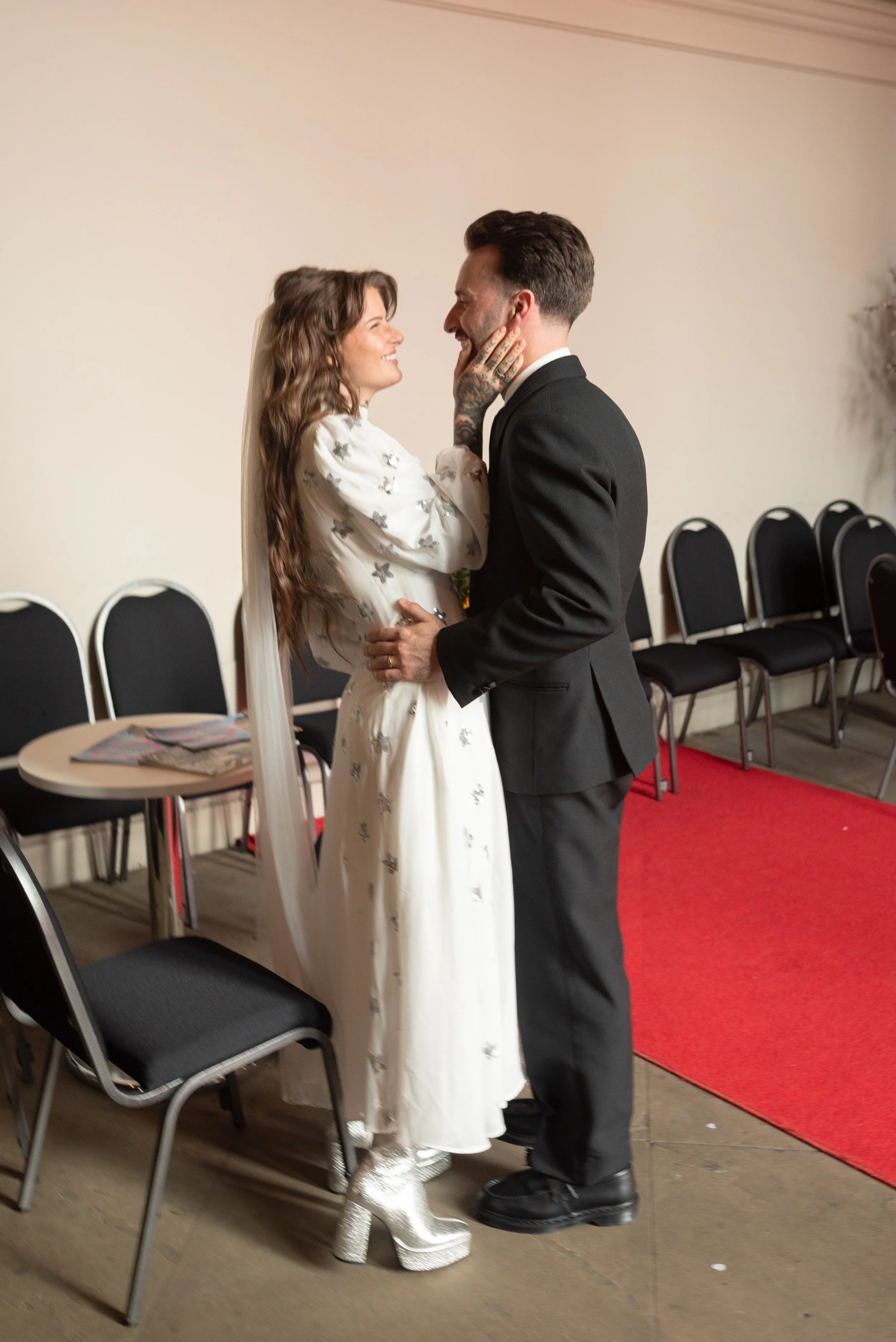 A couple is standing close and smiling at each other. The woman is wearing a white dress with silver details and sparkly silver boots, while the man is in a black suit. The woman has long wavy hair and tattoos on her arm. They are indoors with black 