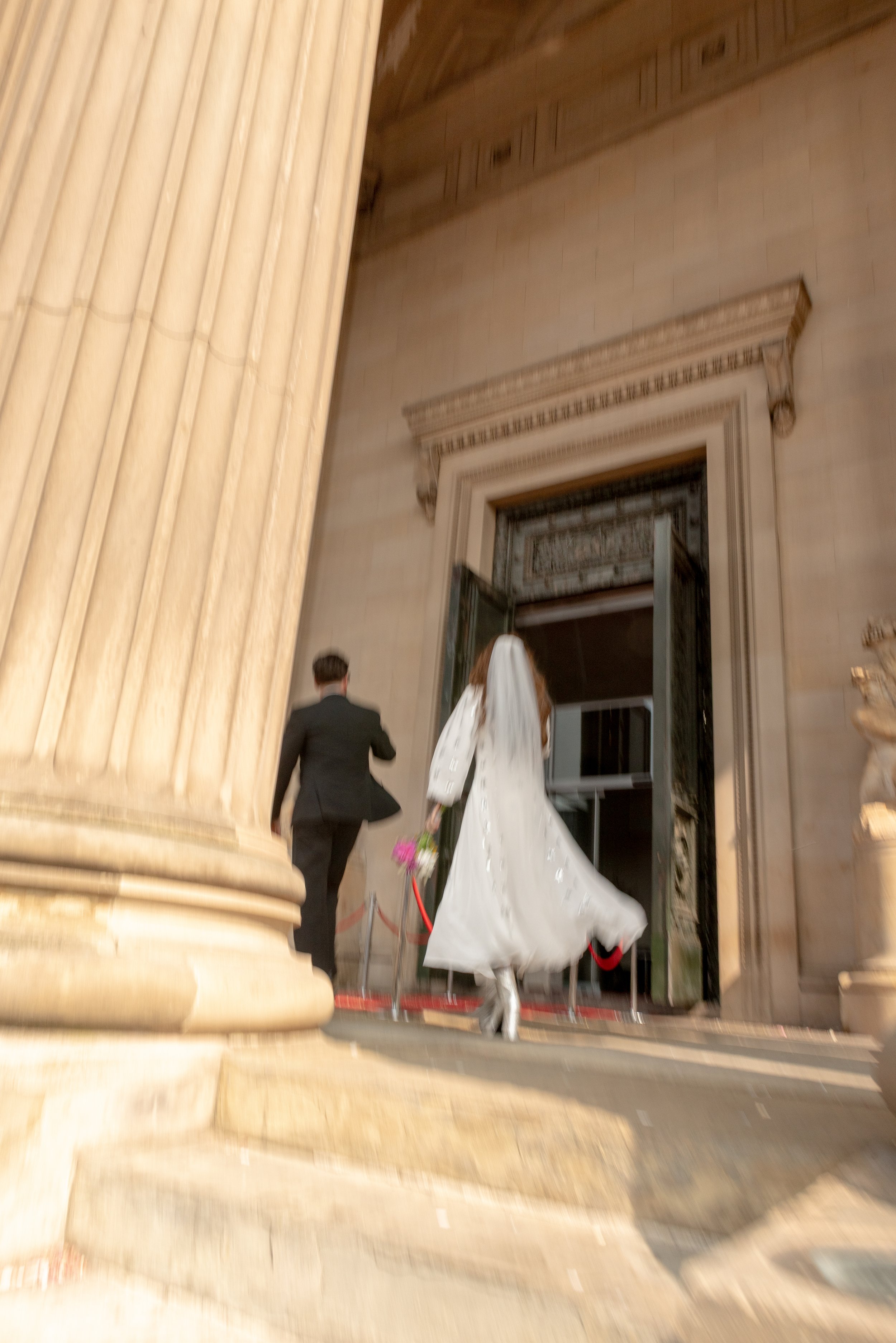 A bride and groom walking into a building, likely a courthouse or historic venue, with large columns and ornate architecture.