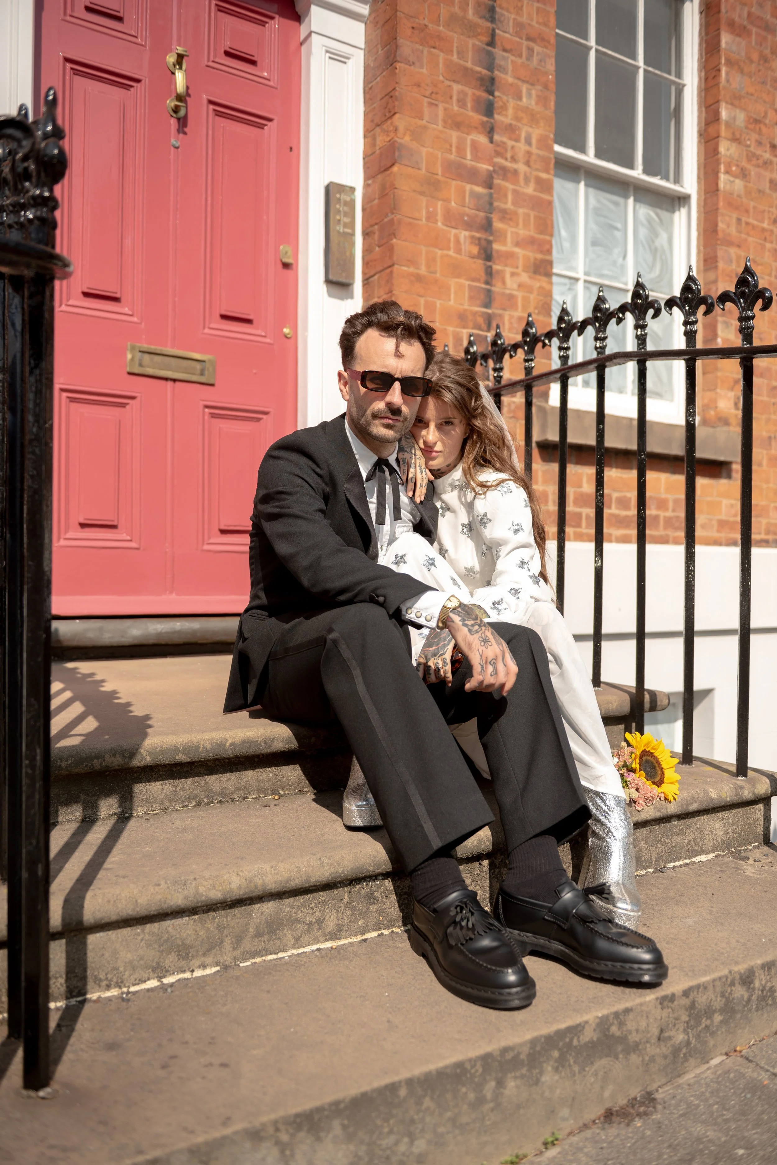 A man and woman sitting on the steps outside a house with a pink door. The man is wearing a black suit, sunglasses, and has tattoos on his hand. The woman is wearing a white dress with silver boots, and has long, wavy hair. There is a sunflower and s