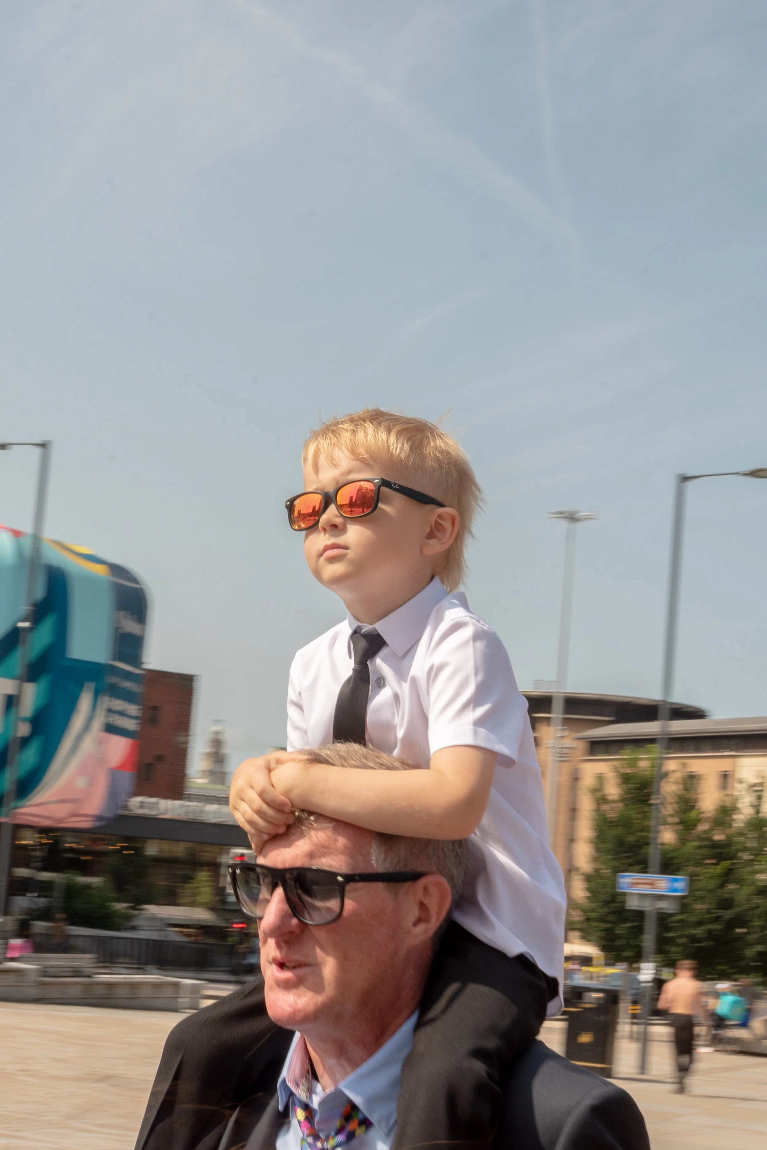 A young boy wearing sunglasses, a white shirt, and a black tie sitting on an older man's shoulders during daytime in an urban area, with buildings, trees, and street signs in the background.