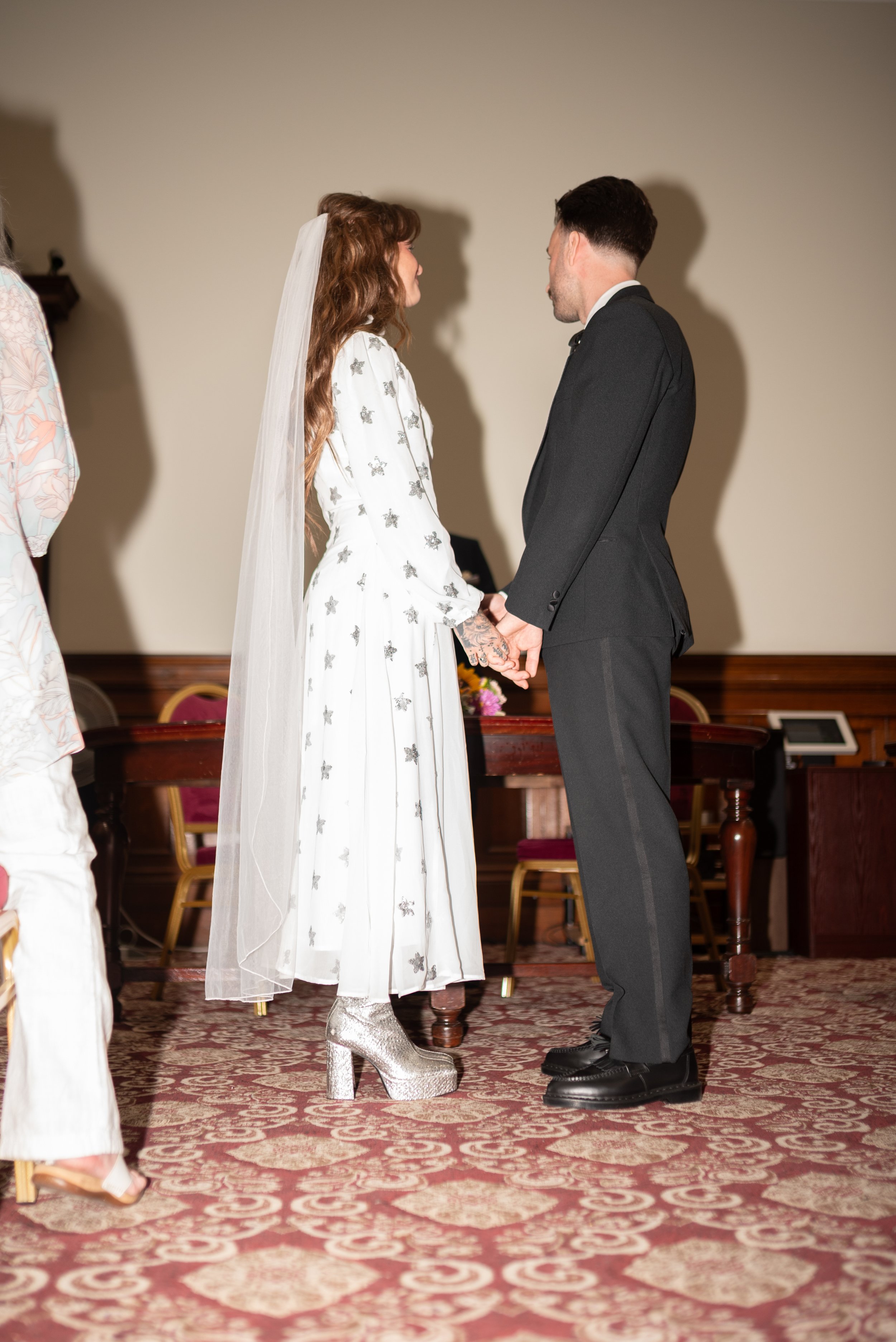 A couple standing face to face holding hands during a wedding ceremony, with a woman in a white wedding dress with metallic high-heeled boots and a veil, and a man in a black suit with black boots, in a room with a patterned carpet and chairs in the 