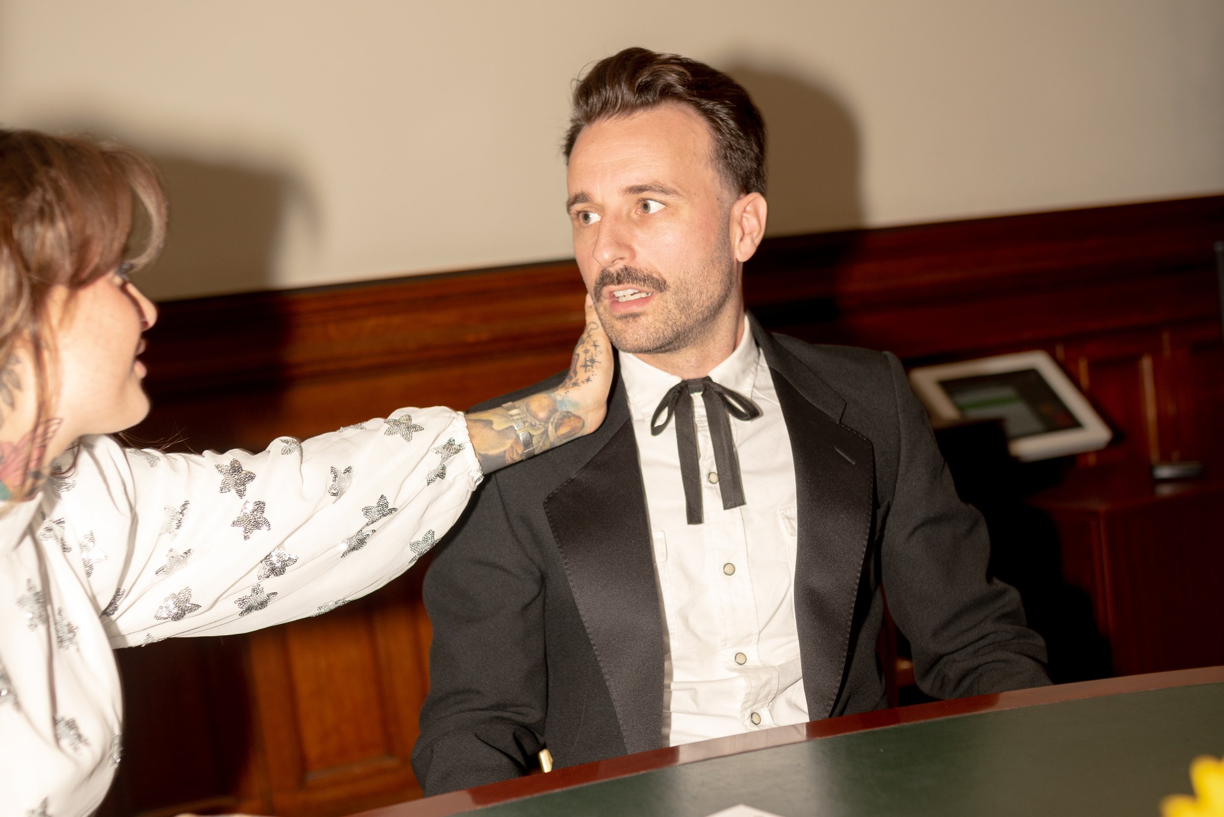 A man in a black tuxedo sitting at a card table, being touched by a woman with tattoos on her arm and hand, who is reaching out to touch his face.
