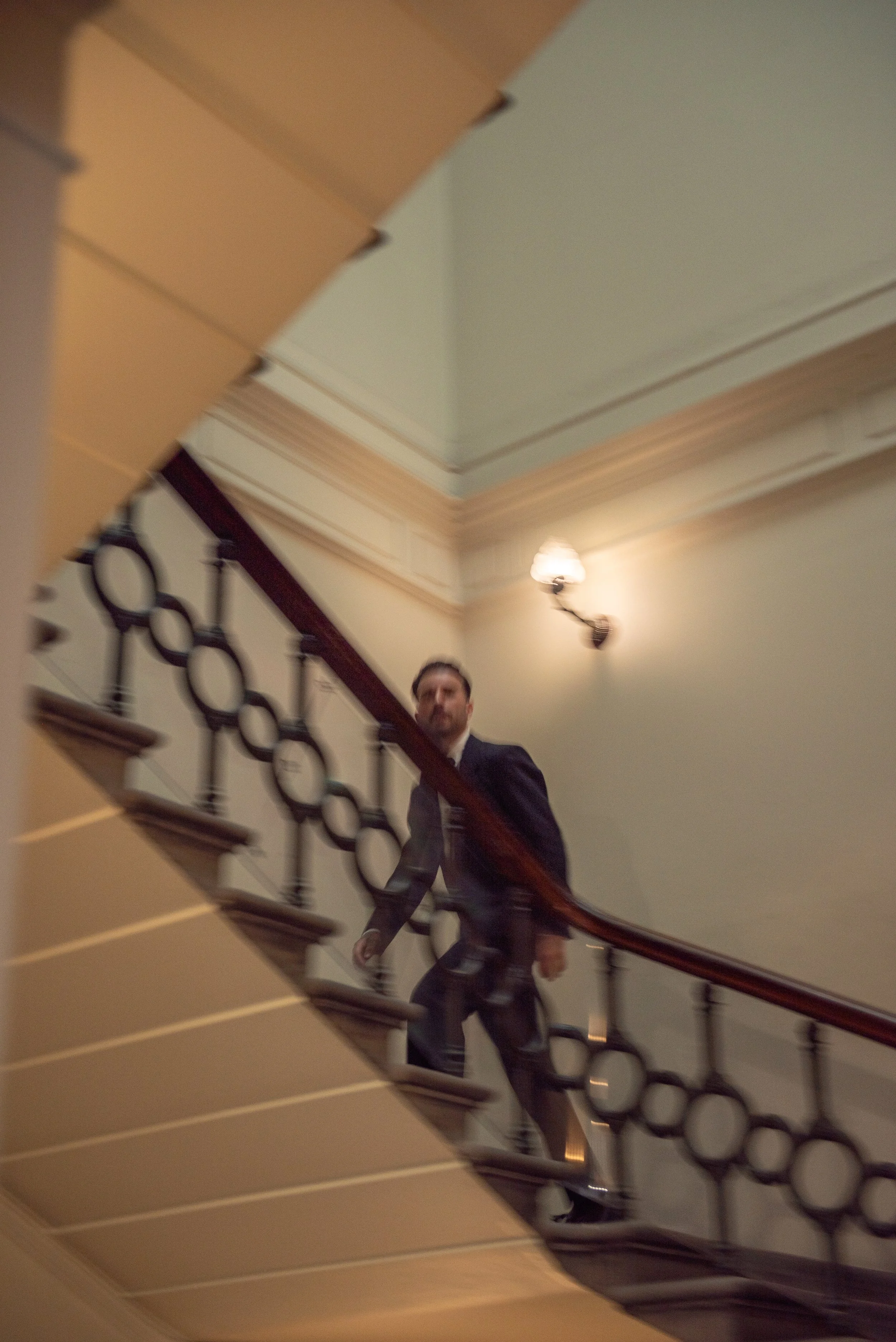 A man in a suit walking up a stairway with a decorative black metal railing, with a wall-mounted light fixture and ceiling molding visible.