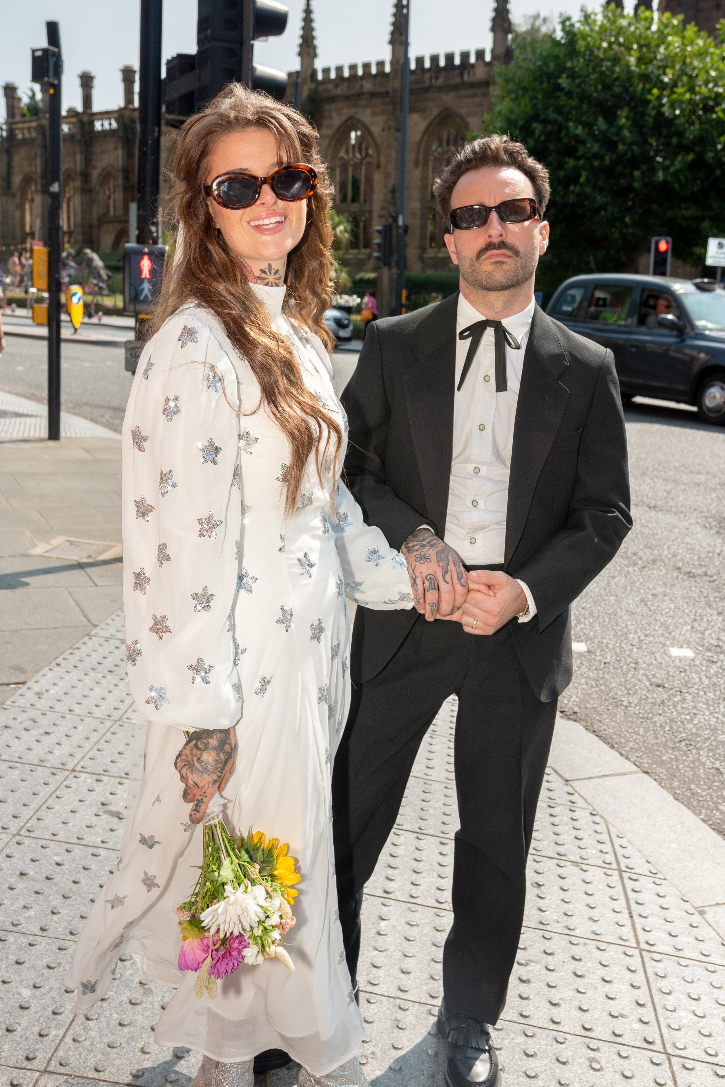 A woman in a white dress with star patterns and sunglasses holding a bouquet of flowers, and a man in a black suit with sunglasses holding her hand, standing on a city sidewalk with traffic and historic buildings in the background.