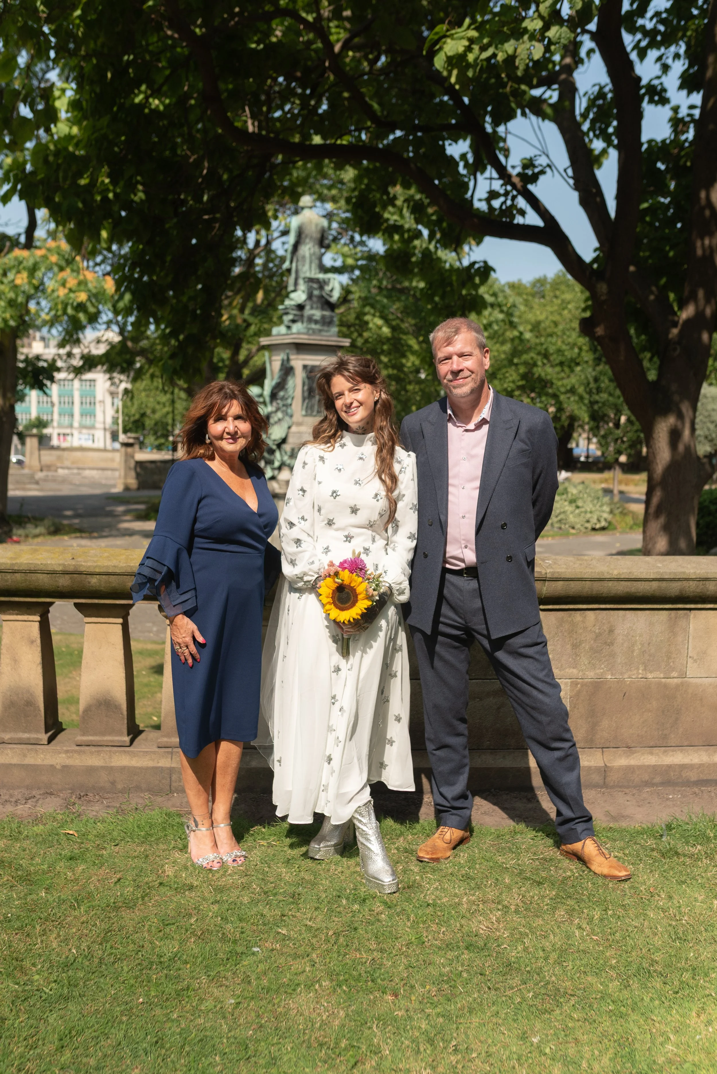 A woman in a white dress holding a sunflower bouquet, flanked by a woman in a blue dress and a man in a gray suit, outdoors under trees, with a monument in the background.