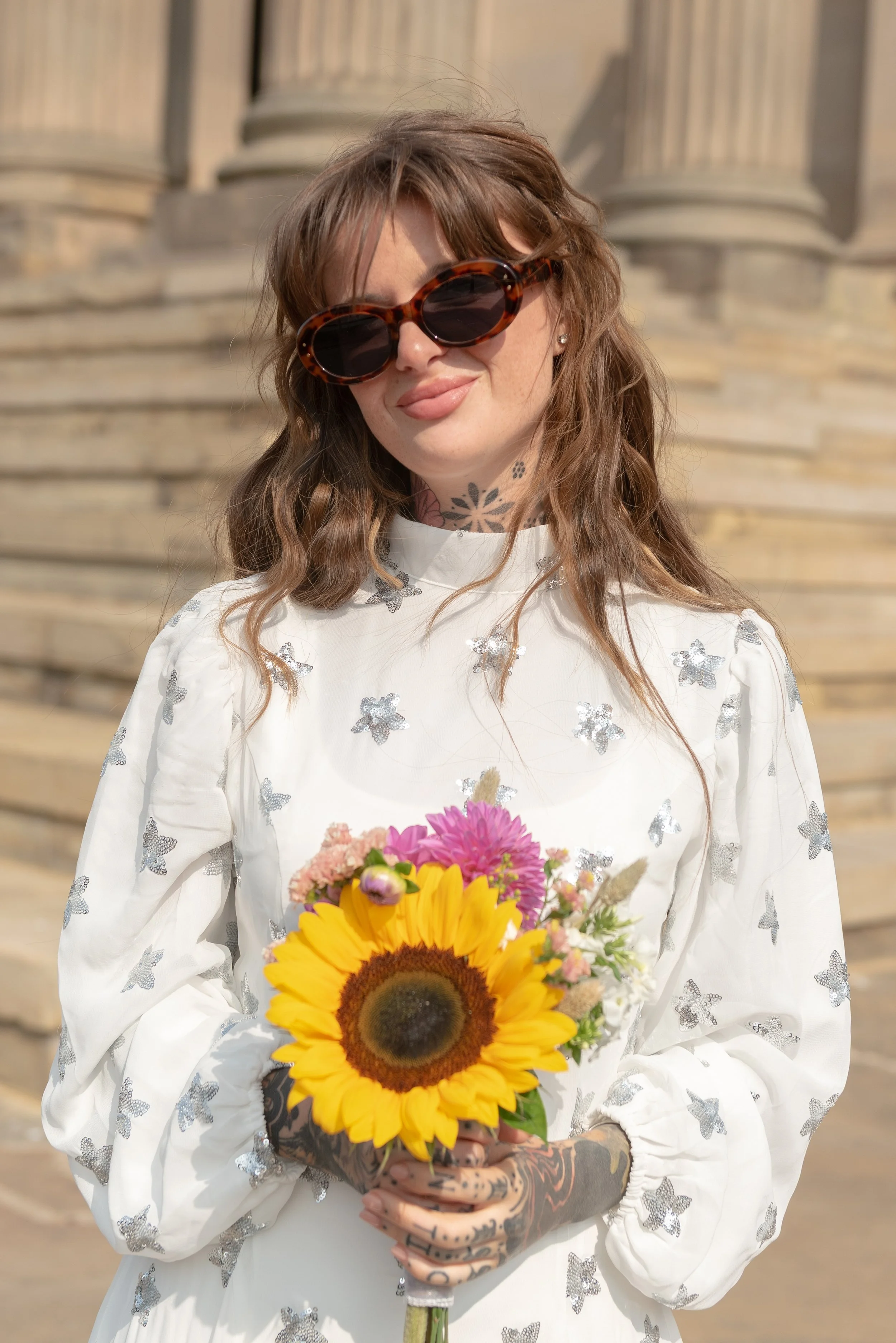 A woman with long wavy hair, wearing large tortoiseshell sunglasses and a white dress with sequined stars, holding a bouquet of colorful flowers including a sunflower and pink blossoms, standing outdoors in front of stone steps.