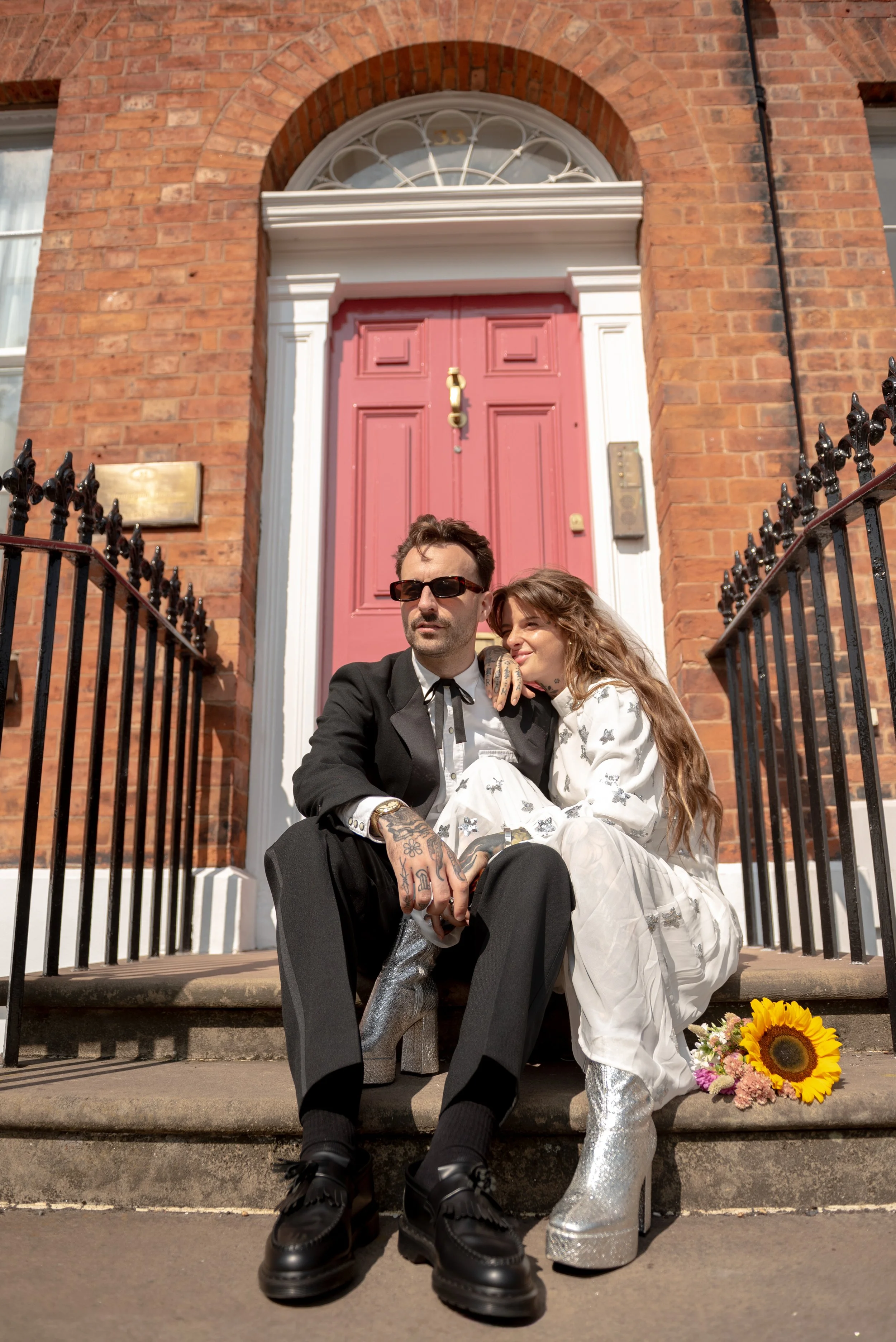 A man and woman sitting on front steps in front of a red brick building with a pink door. The man wears a black suit, sunglasses, and black shoes, with tattoos on his hands. The woman wears a white dress with butterfly patterns and silver high-heeled