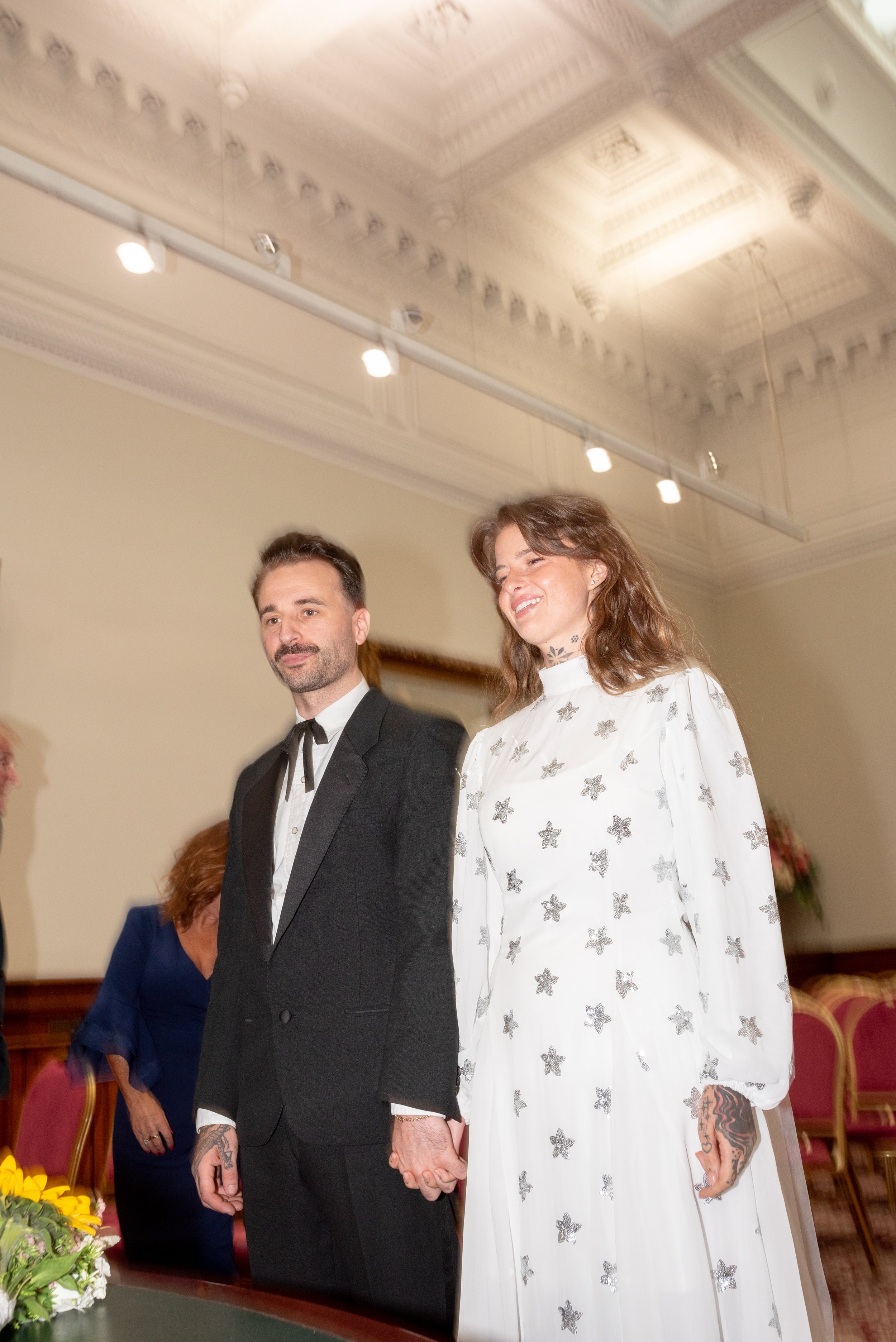 A couple holding hands during a formal wedding ceremony, with the woman smiling and dressed in a white dress with butterfly patterns and the man in a black suit with a bow tie, in a decorated indoor setting.