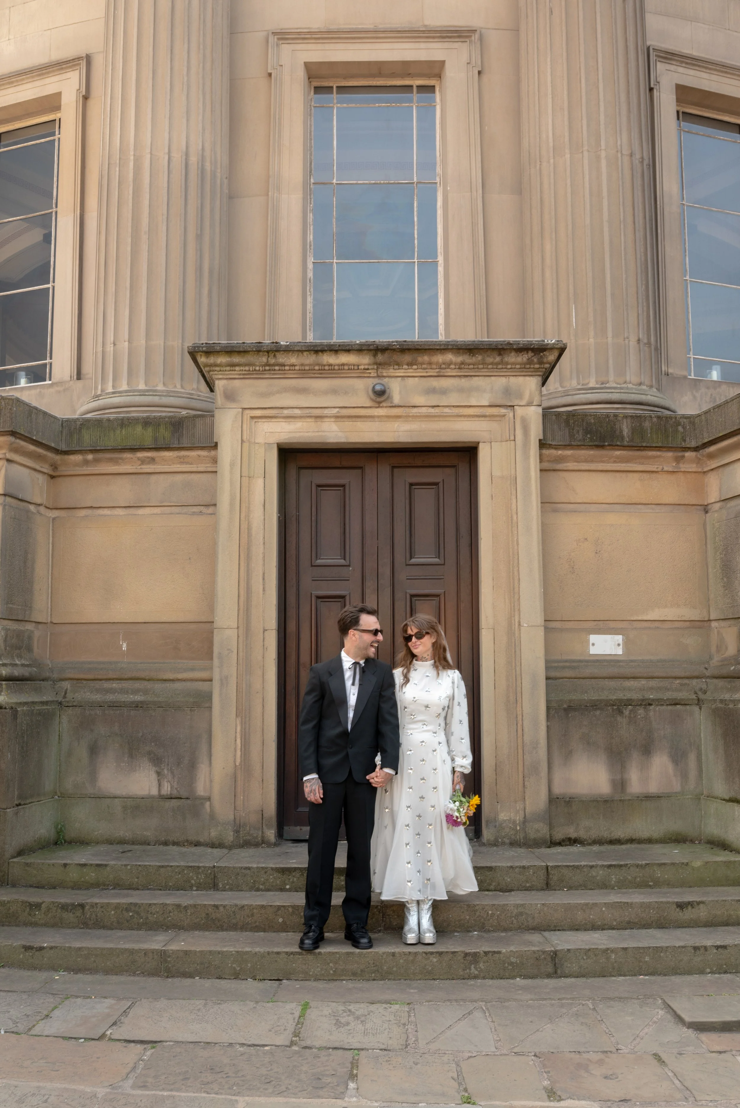 A couple dressed in wedding attire standing on the steps in front of a large historic building with tall windows and columns, holding hands and smiling at each other.