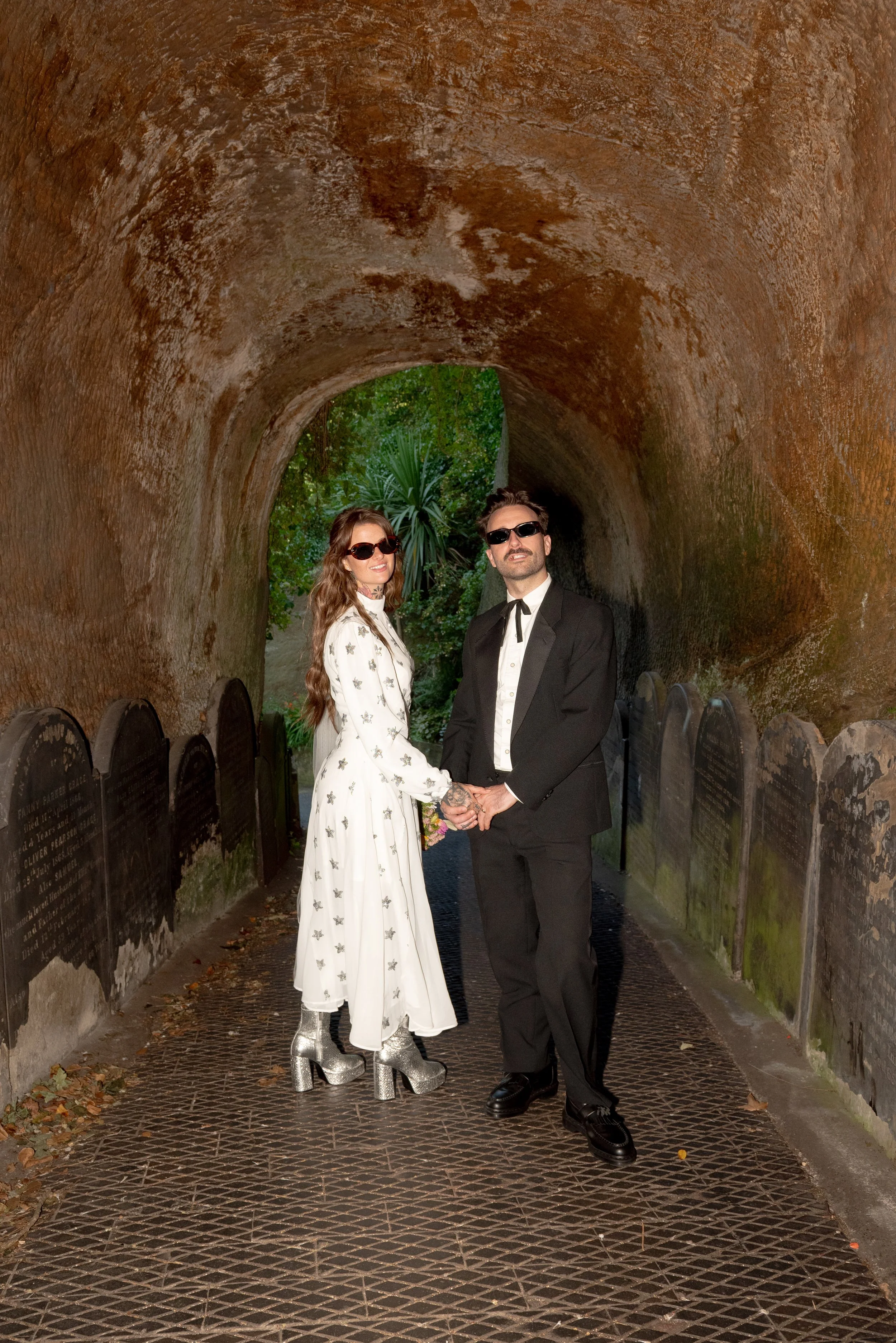 A couple dressed in vintage style standing hand in hand in front of a rock tunnel with lush greenery, wearing sunglasses and fashionable clothes.