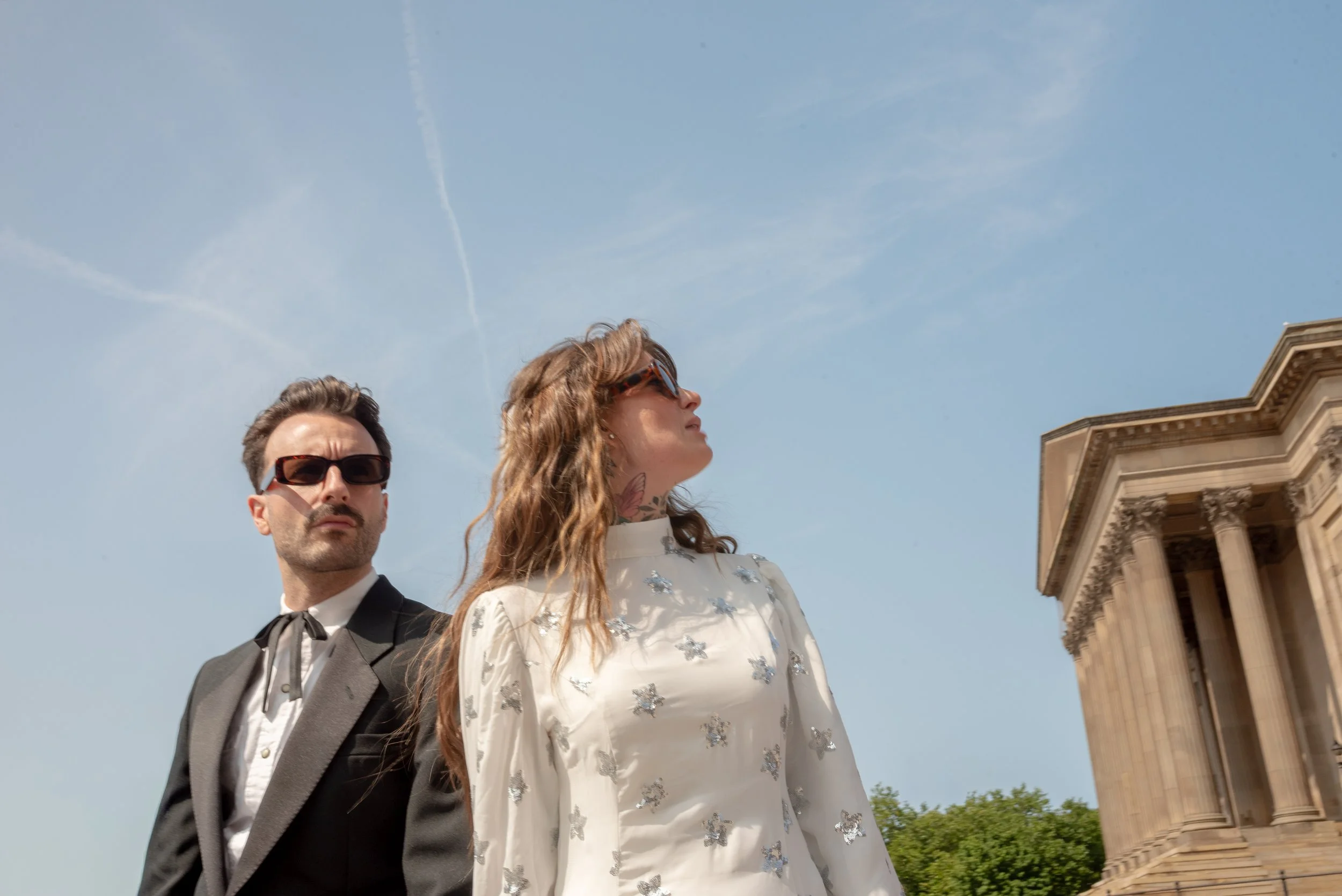 A man in a tuxedo and a woman in a white dress with silver embellishments standing outdoors near a historic building with columns, under a blue sky with some clouds and contrails.