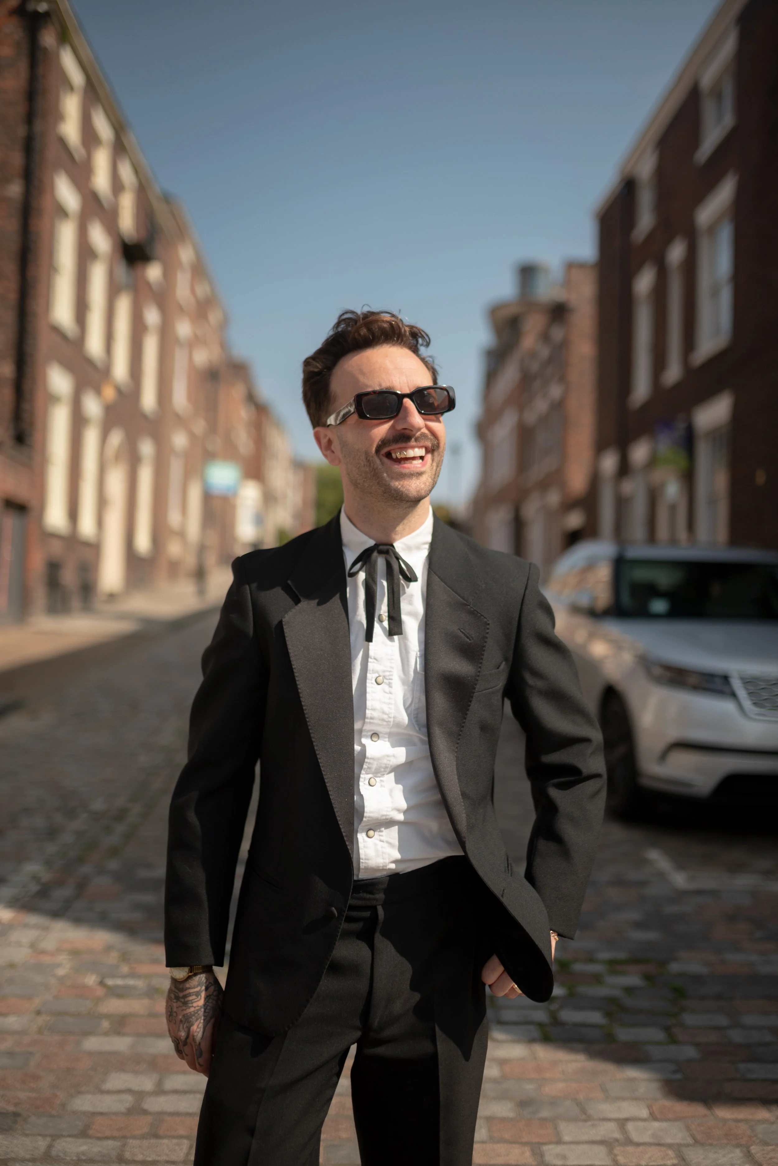 A man in a black suit, white shirt, and black tie with a bow, wearing sunglasses, is smiling and walking on a cobblestone street with brick buildings and a white car in the background.