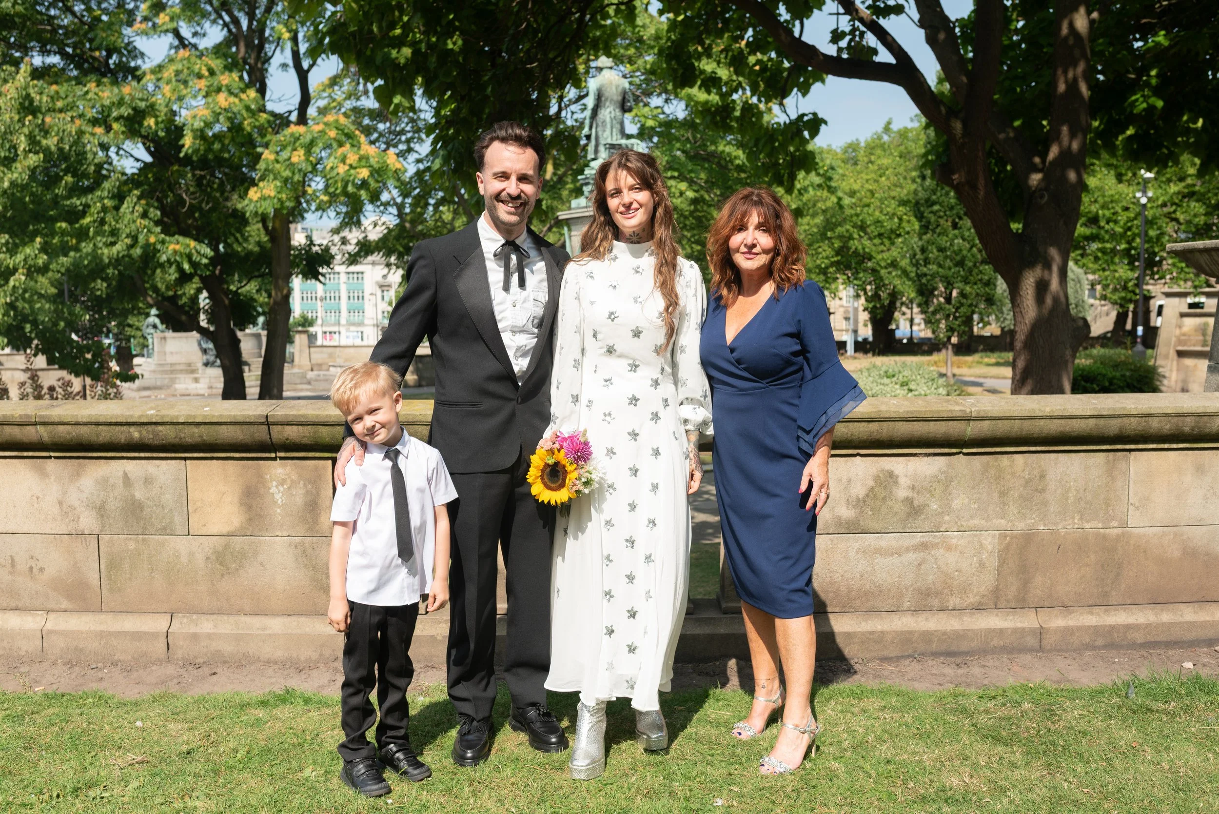 Family standing outdoors in a park, celebrating a special occasion, with trees and a monument in the background.