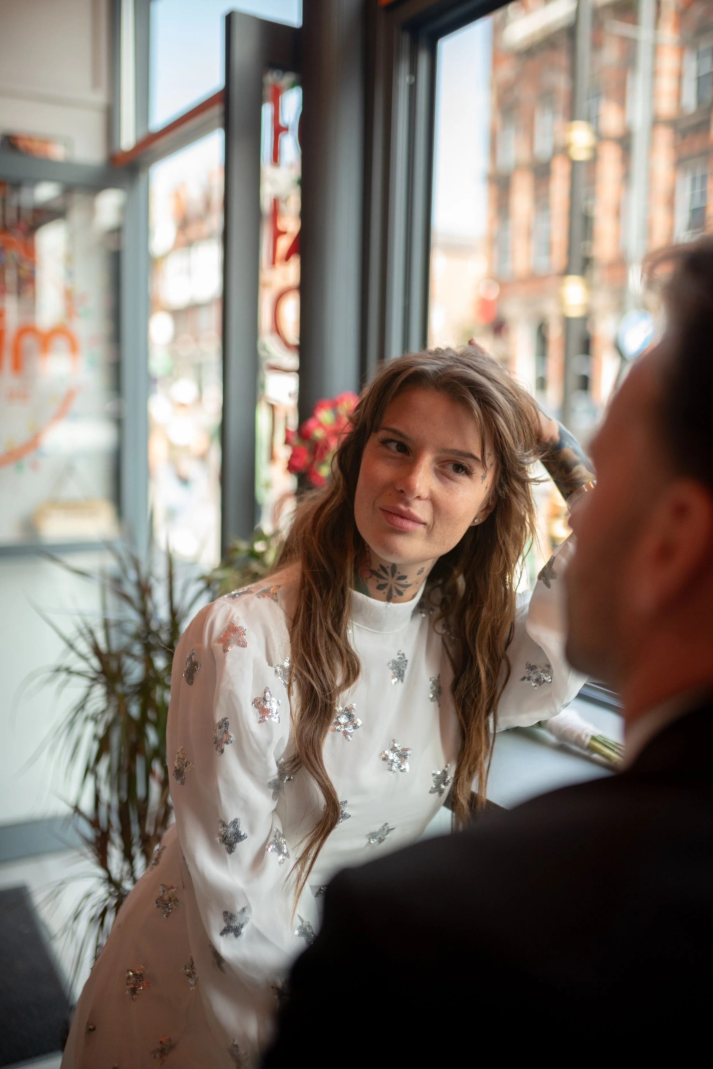 Woman with long brown hair and tattoos on her neck and arms, wearing a white dress with silver butterflies, sitting in a cafe by a window, looking at a man with short dark hair.