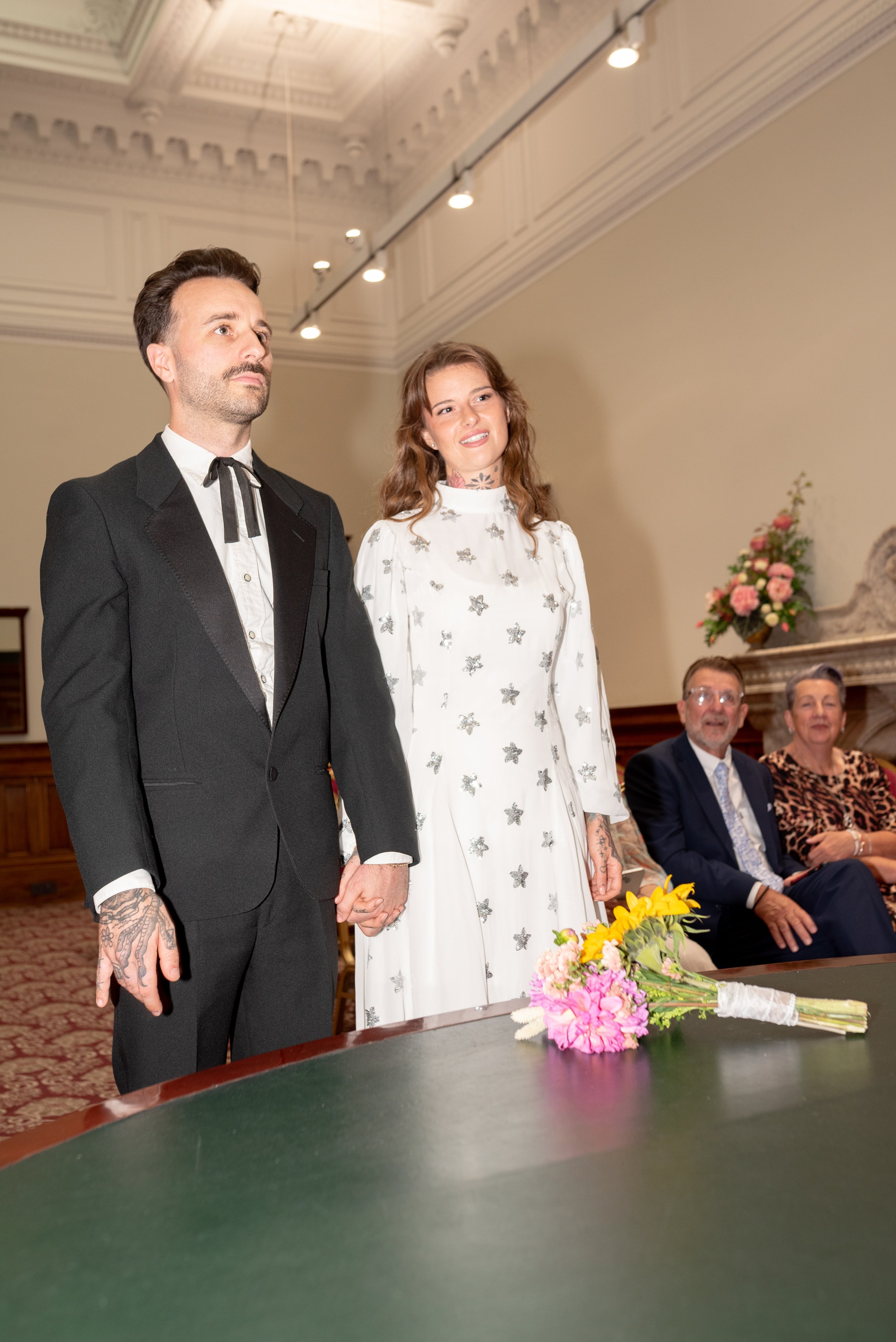 A couple holding hands standing at a wedding ceremony, with two people seated behind them, in a decorated indoor setting.