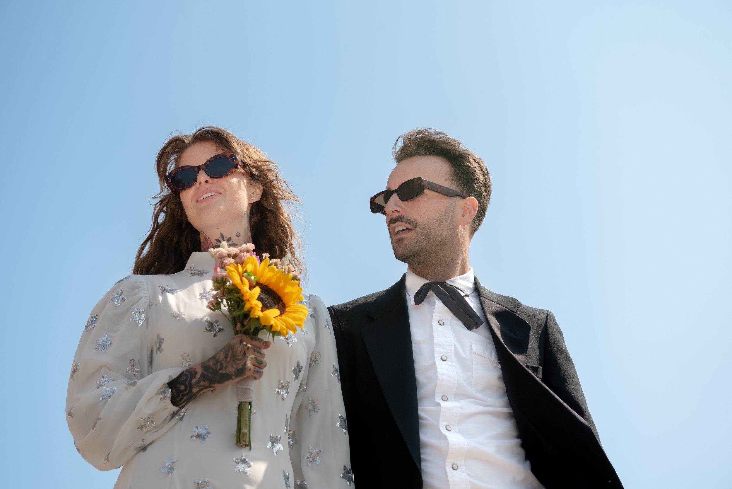 A woman holding a sunflower bouquet wearing sunglasses and a white embroidered dress, standing next to a man in a black suit with sunglasses, against a clear blue sky.