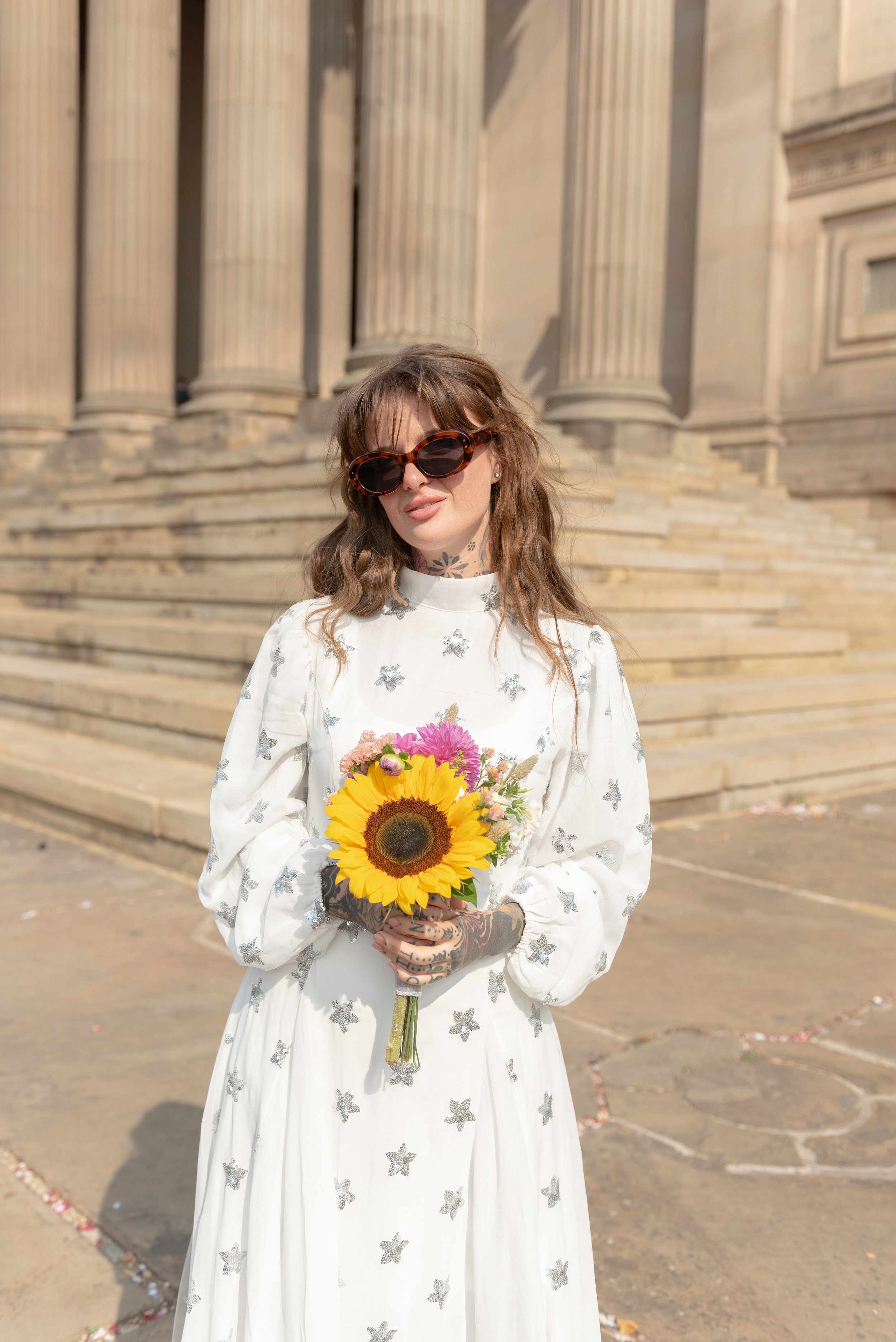 A woman with long brown hair, wearing large sunglasses and a white dress with butterfly patterns, holds a bouquet of colorful flowers, including a sunflower, in front of a grand stone building with columns and steps.