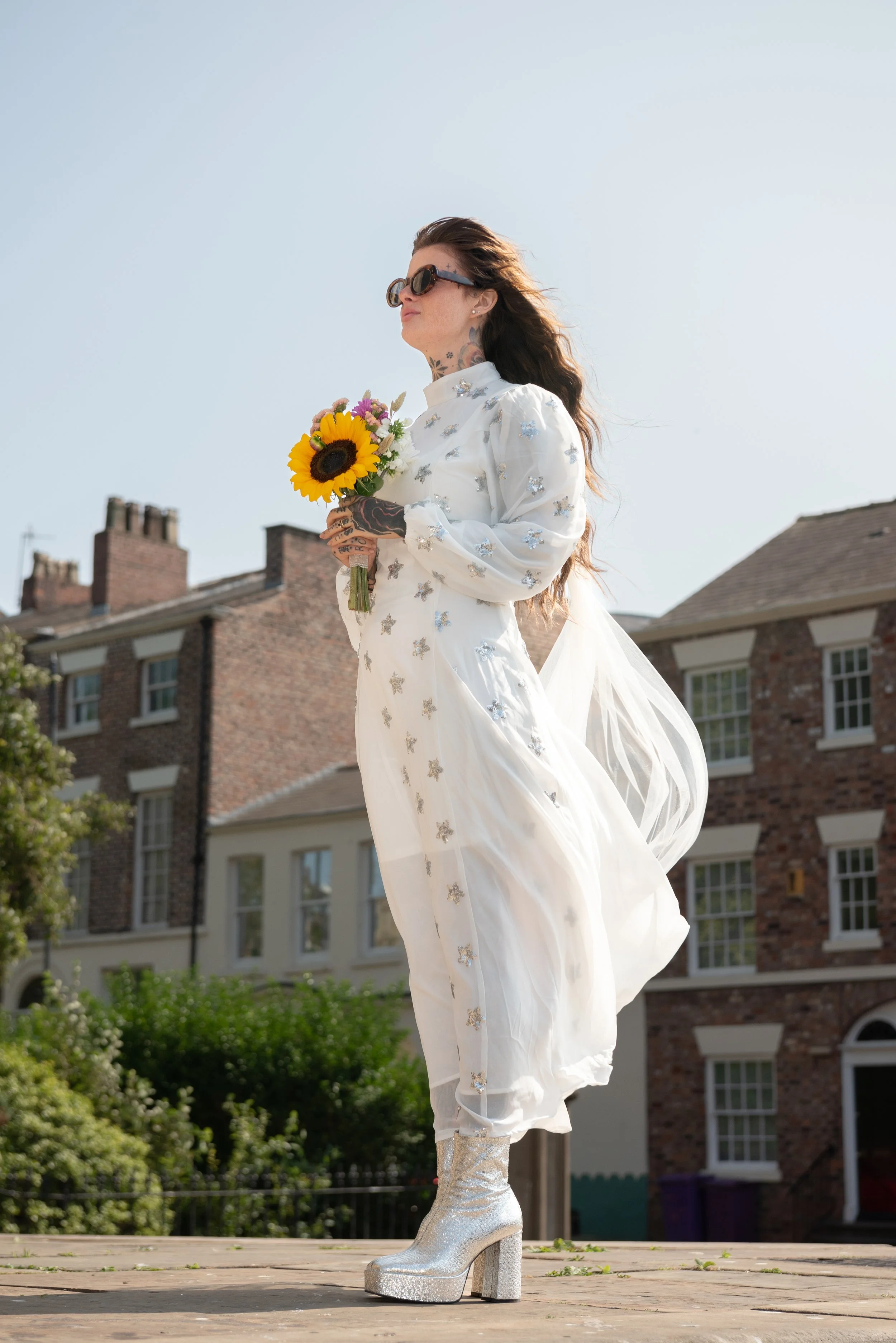 Woman standing on a wooden surface outdoors, holding a small bouquet of flowers, with residential buildings in the background, wearing a long white dress and silver glittery platform boots, sunglasses, and having long hair.