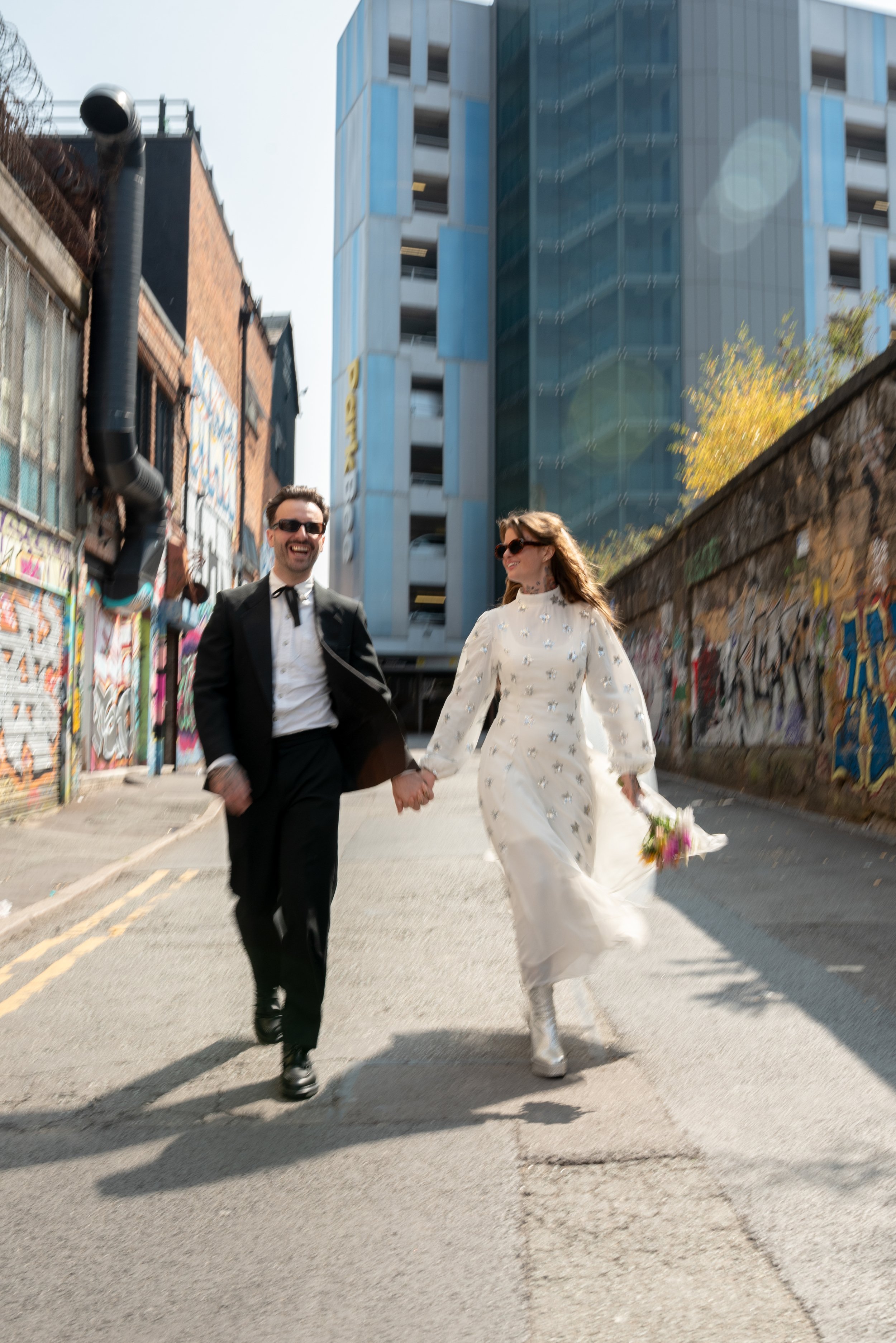 A couple dressed in wedding attire, holding hands and walking down an urban street with graffiti-covered walls and modern buildings.