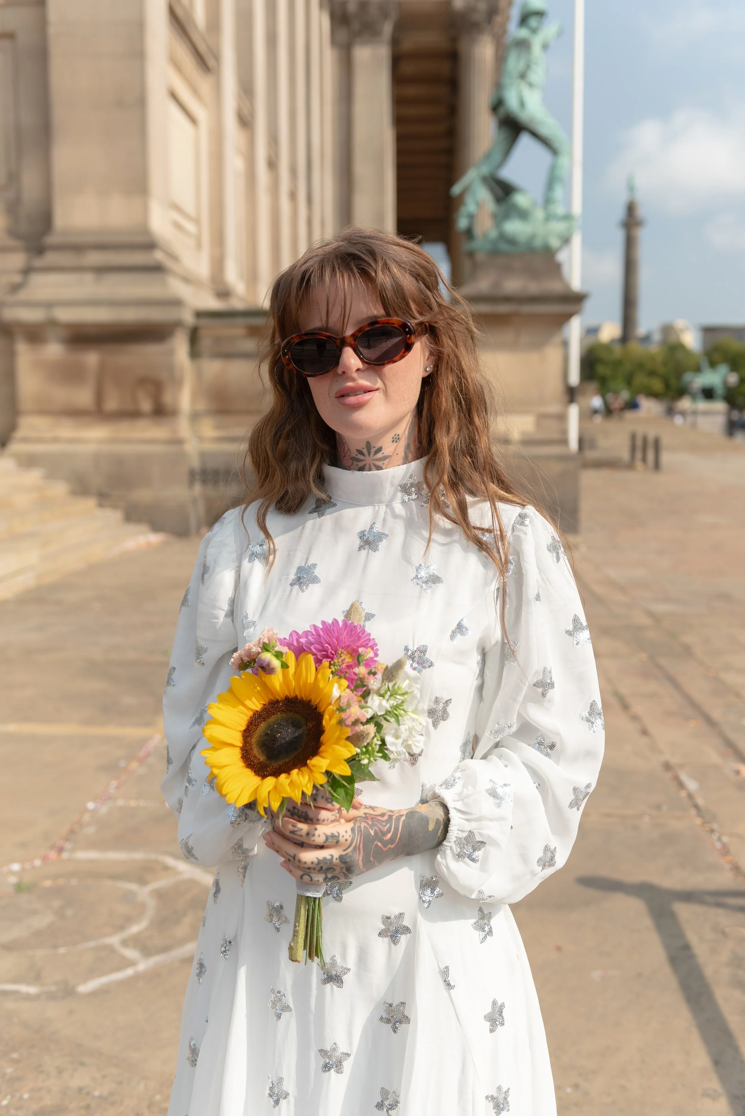 A woman with wavy brown hair wearing large tortoiseshell sunglasses and a white dress with silver butterfly patterns. She holds a bouquet of sunflowers and pink and white flowers, standing outdoors near a historical building with sculptures and colum