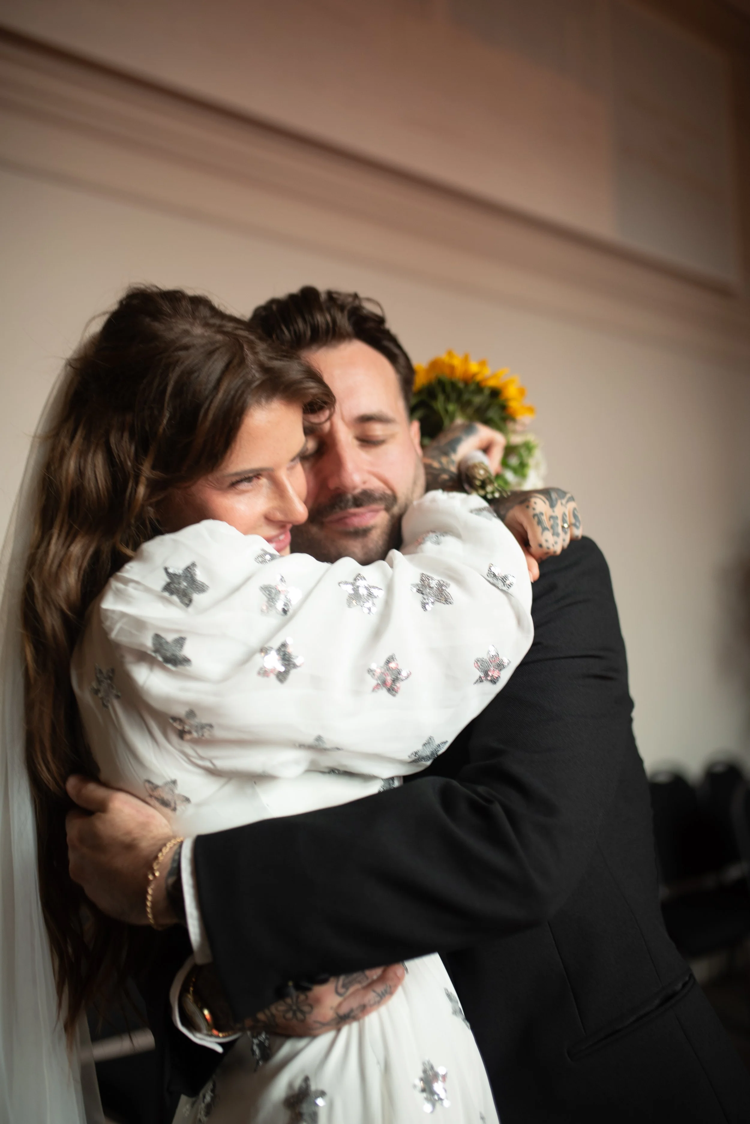 A man and woman hugging, the woman wearing a white dress with silver star embellishments, and the woman holding a bouquet of sunflowers behind him.