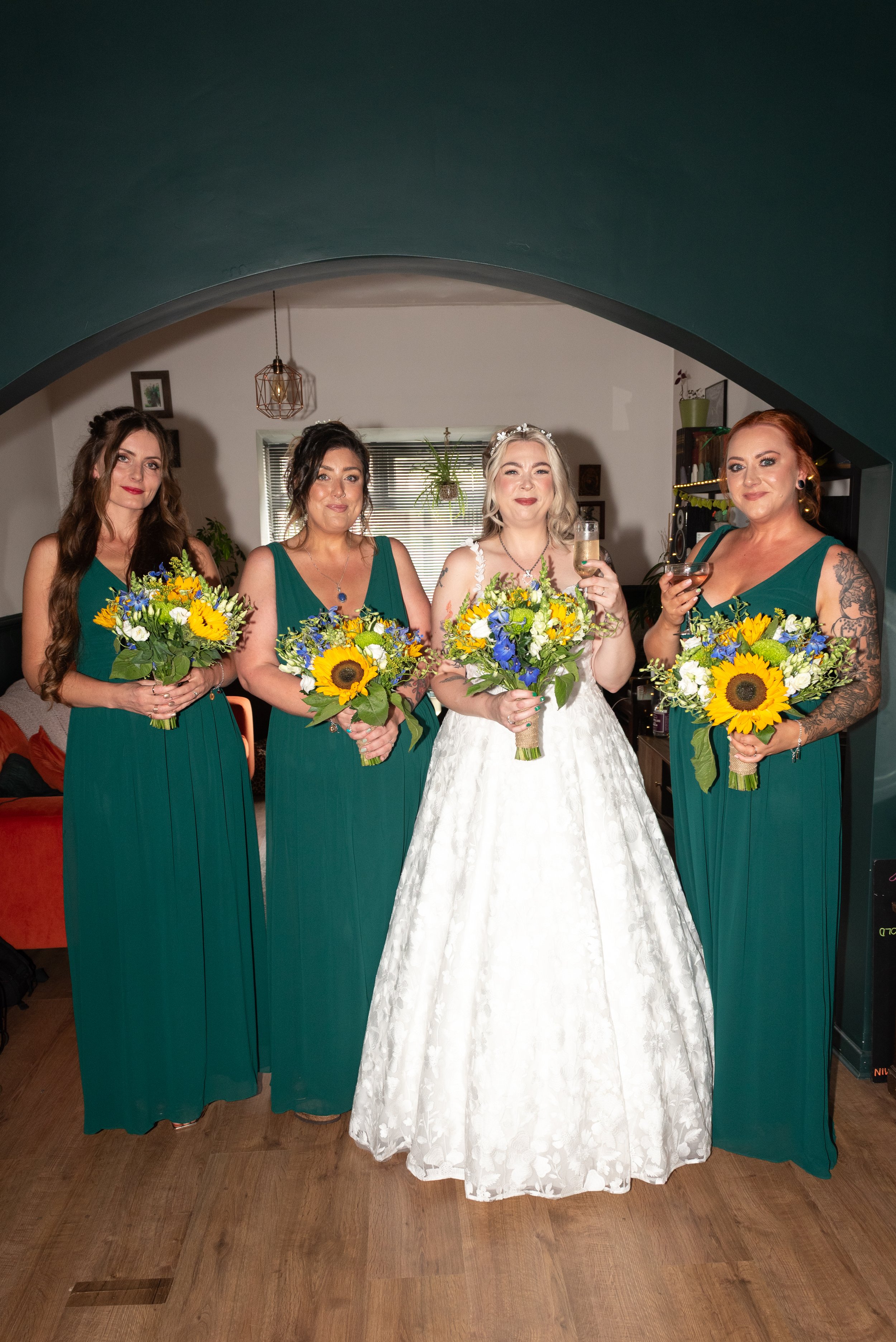 A bride in a white wedding gown and three bridesmaids in green dresses holding bouquets of yellow, white, and purple flowers, standing indoors under an arched ceiling, with two women holding glasses of champagne.