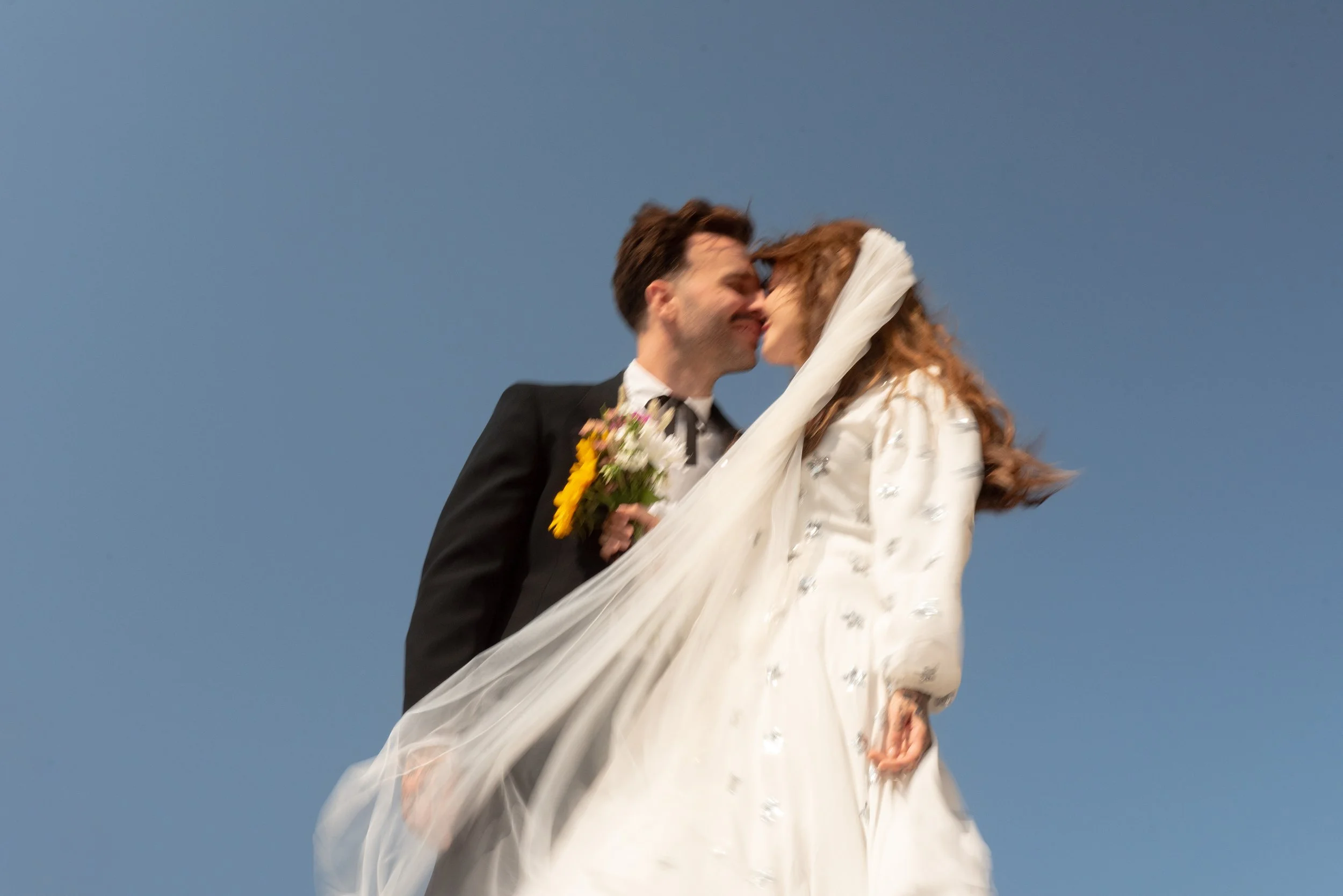 A bride and groom sharing a kiss outdoors against a clear blue sky, with the bride holding a bouquet of flowers.