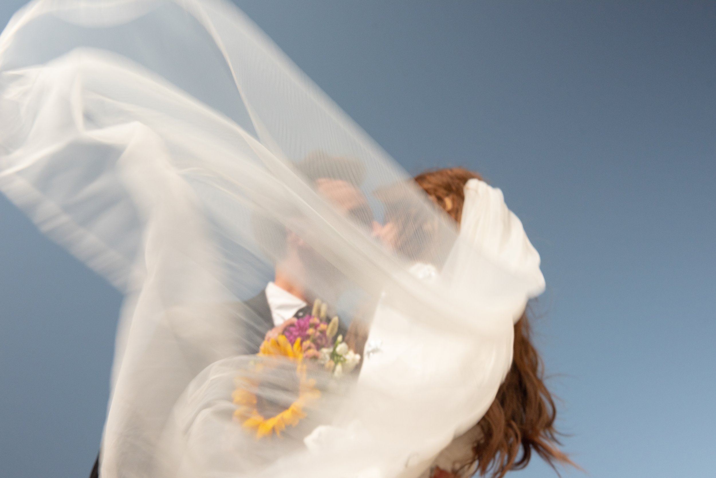 A woman with curly hair holding a bouquet of flowers, partially covered by a flowing white veil against a clear blue sky.