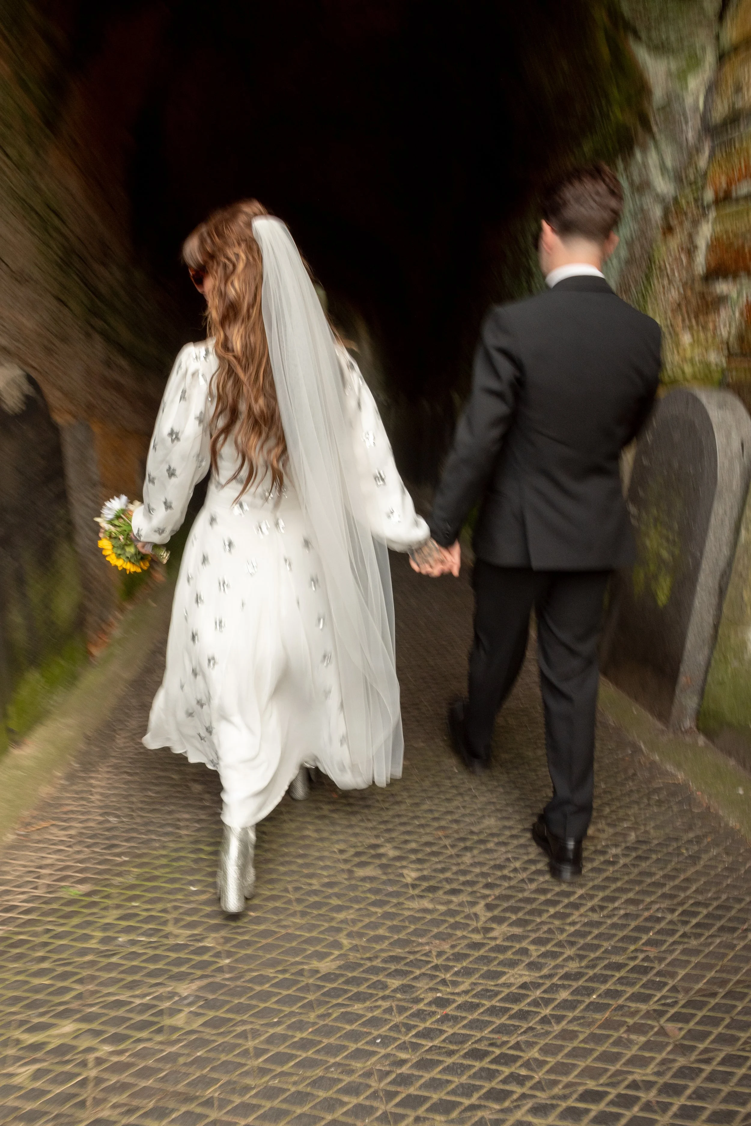 A bride and groom holding hands and walking away through a tunnel or cave, with the bride wearing a white dress, veil, and holding a small bouquet, and the groom in a black suit.