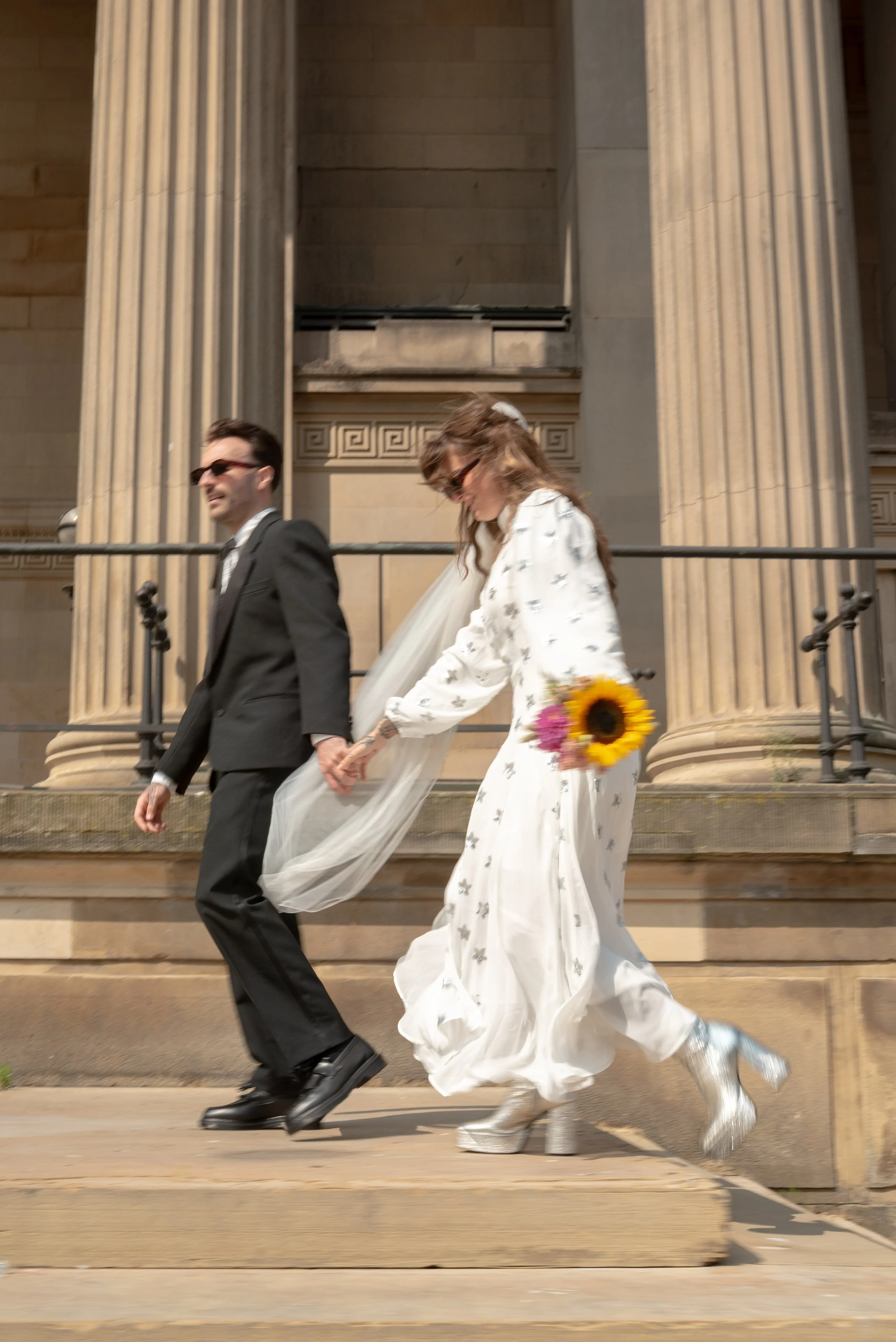 A bride and groom holding hands while walking down steps outside a classical building with large columns. The bride is holding a bouquet of sunflowers and wearing silver boots. The groom is dressed in a black suit and sunglasses.