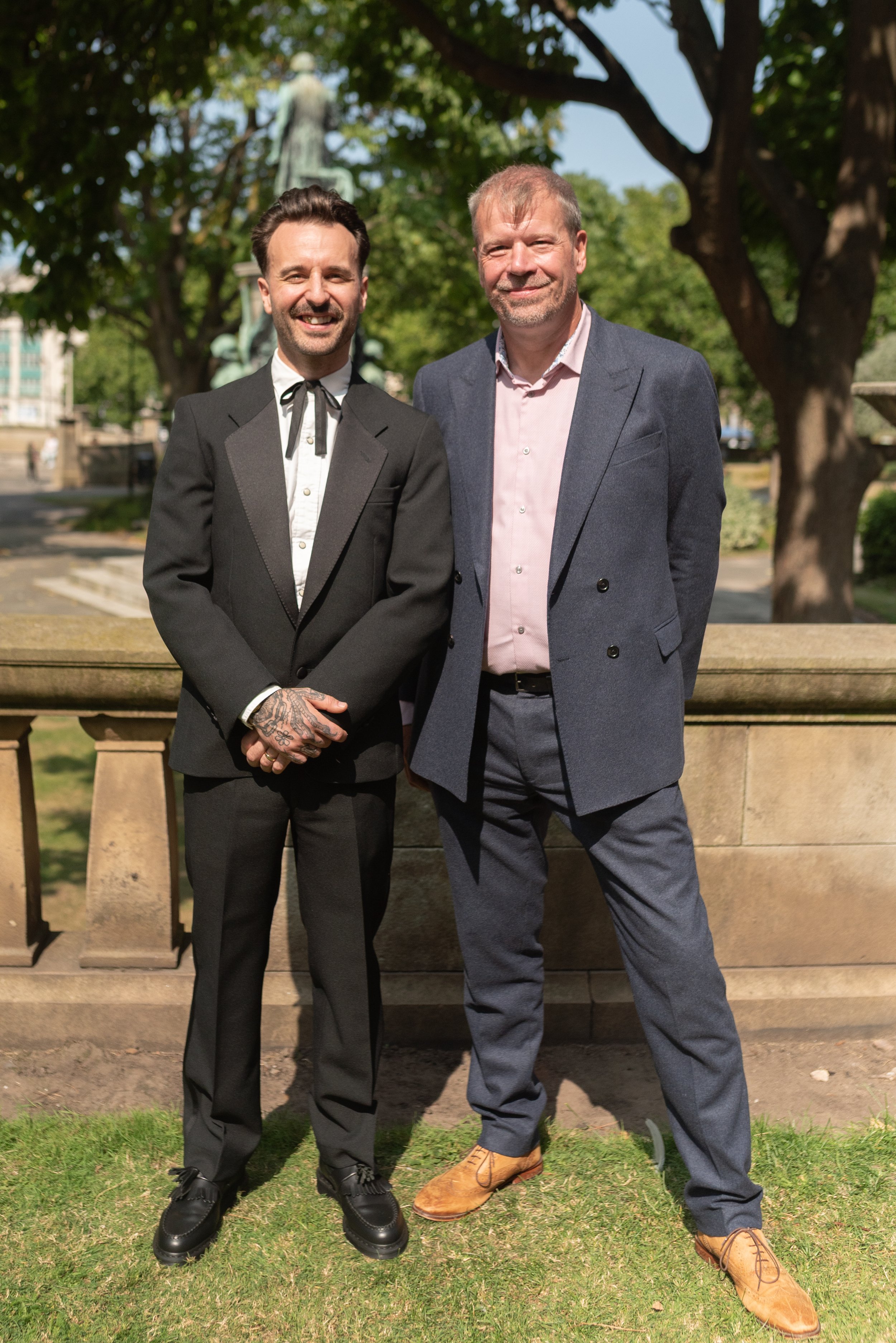 Two well-dressed men standing outdoors in front of a stone railing with a statue and trees in the background. One man is wearing a black suit with a bow tie, and the other is wearing a grey suit with a pink shirt. Both are smiling.