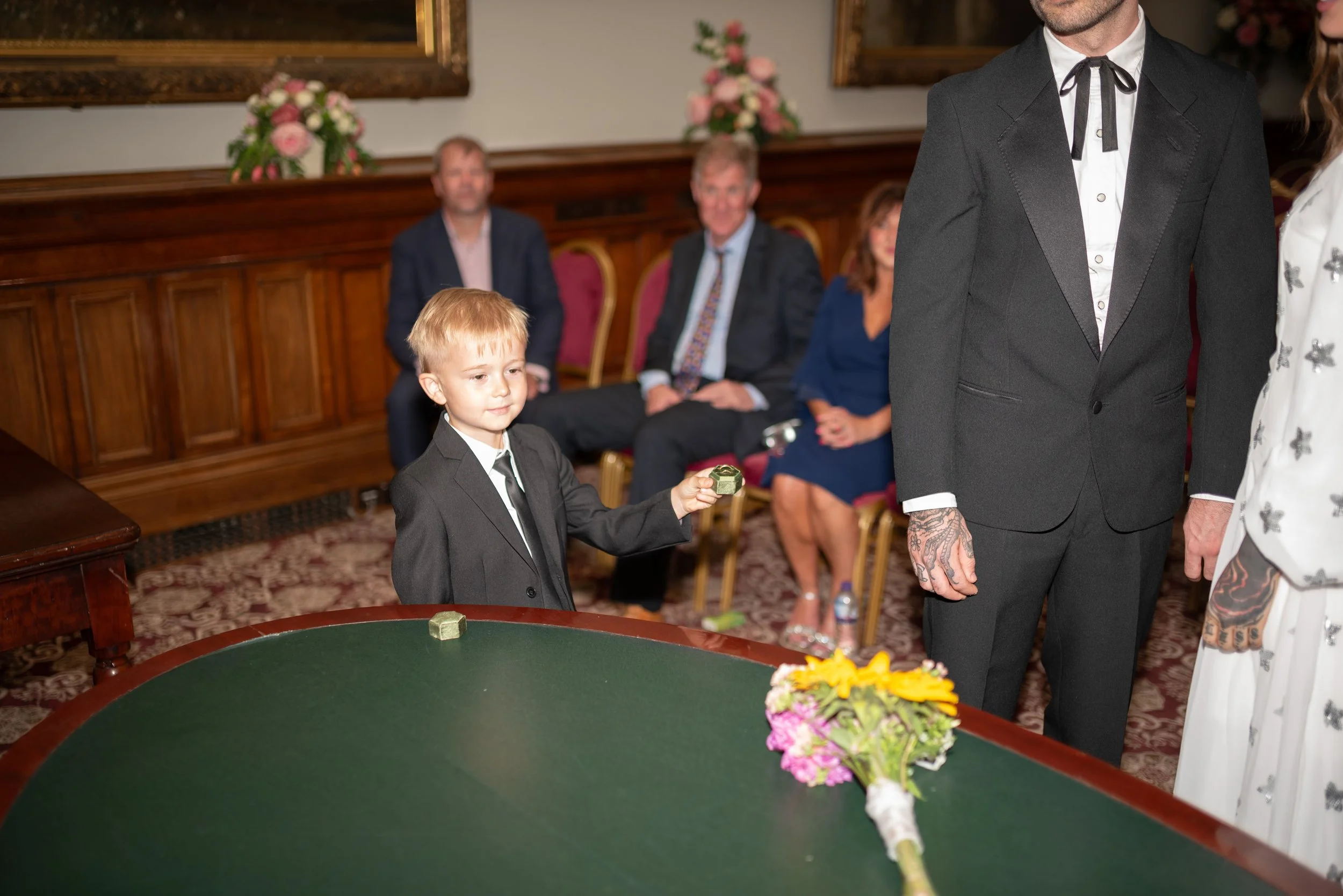 A young boy in a black suit holding an object near a green table, with a bouquet of flowers on it, during a formal event with three people sitting in the background and two people standing to the right.