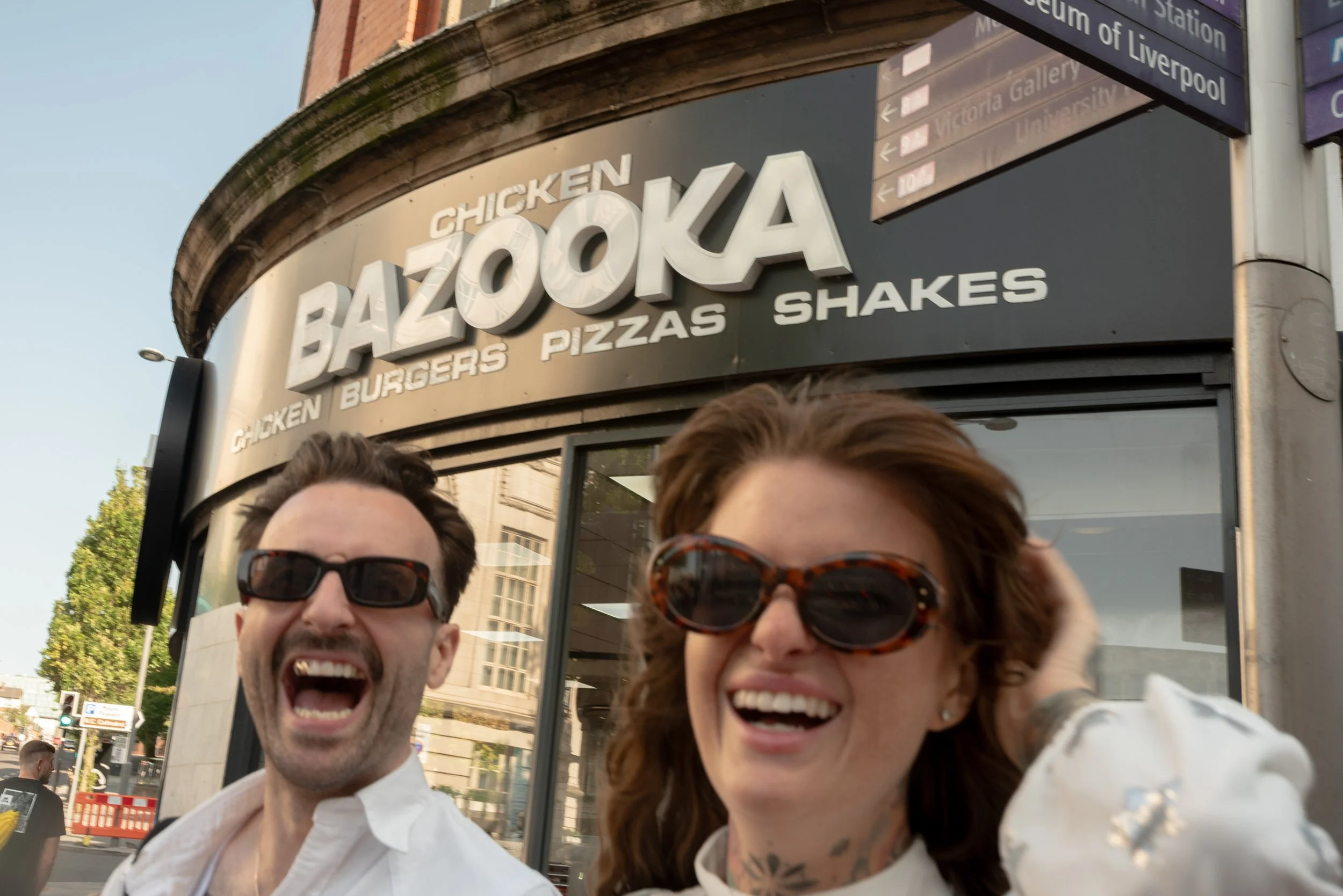 Two people smiling and wearing sunglasses in front of a Bazooka restaurant sign that offers chicken, burgers, pizzas, and shakes.