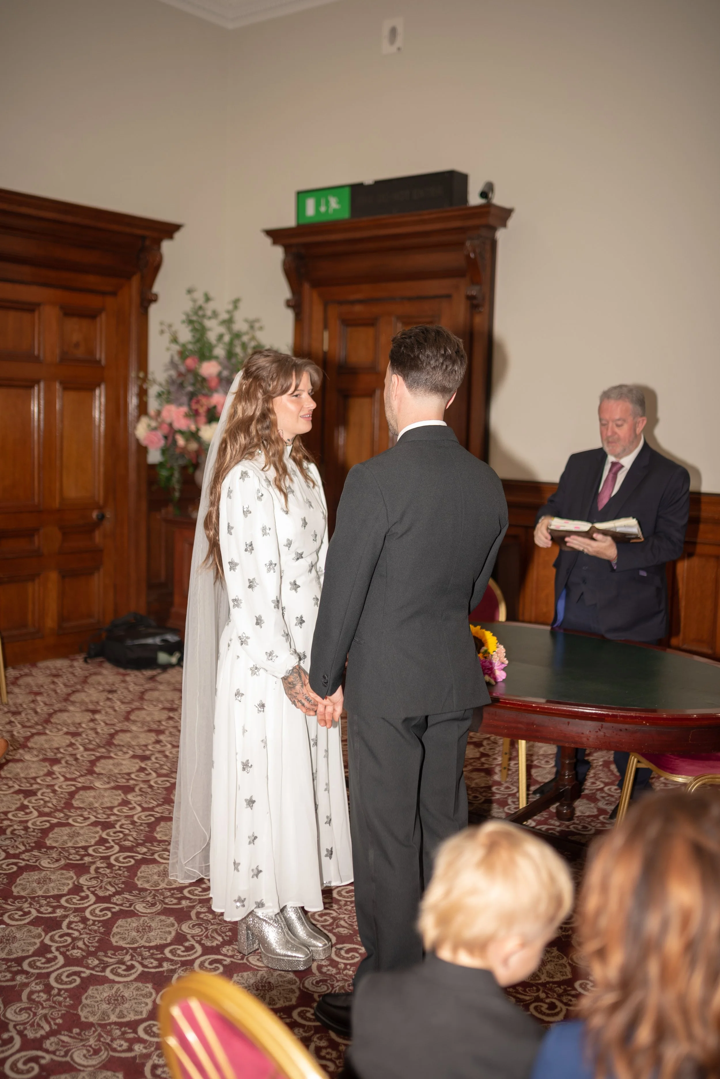 A bride and groom holding hands during their wedding ceremony, facing each other, with an officiant reading in the background, inside a room with wooden paneling and floral decorations.