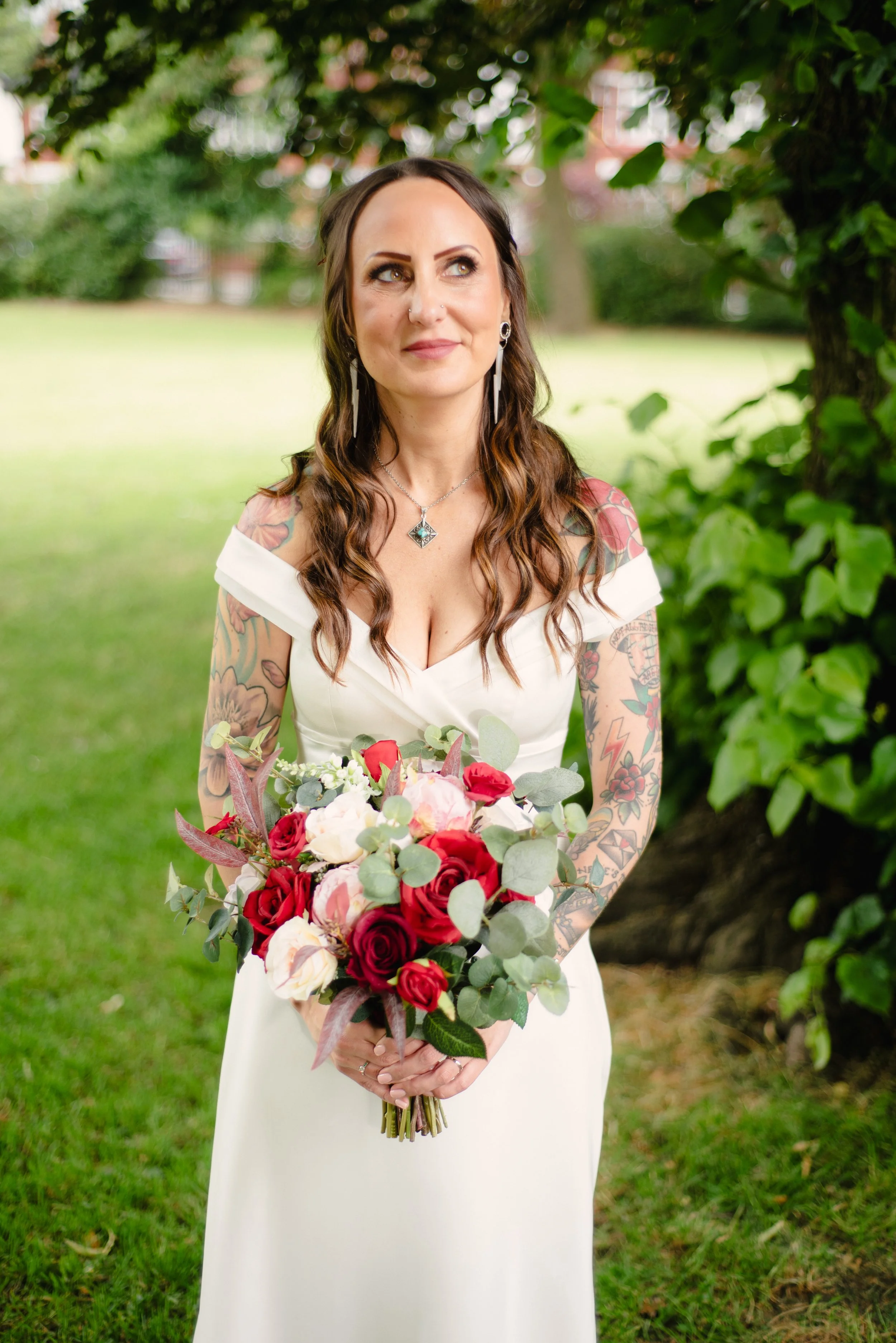 A woman with brown wavy hair and tattoos on her arms, wearing a white dress, holding a bouquet of red and pink roses with green foliage, standing outdoors in a green park with trees.