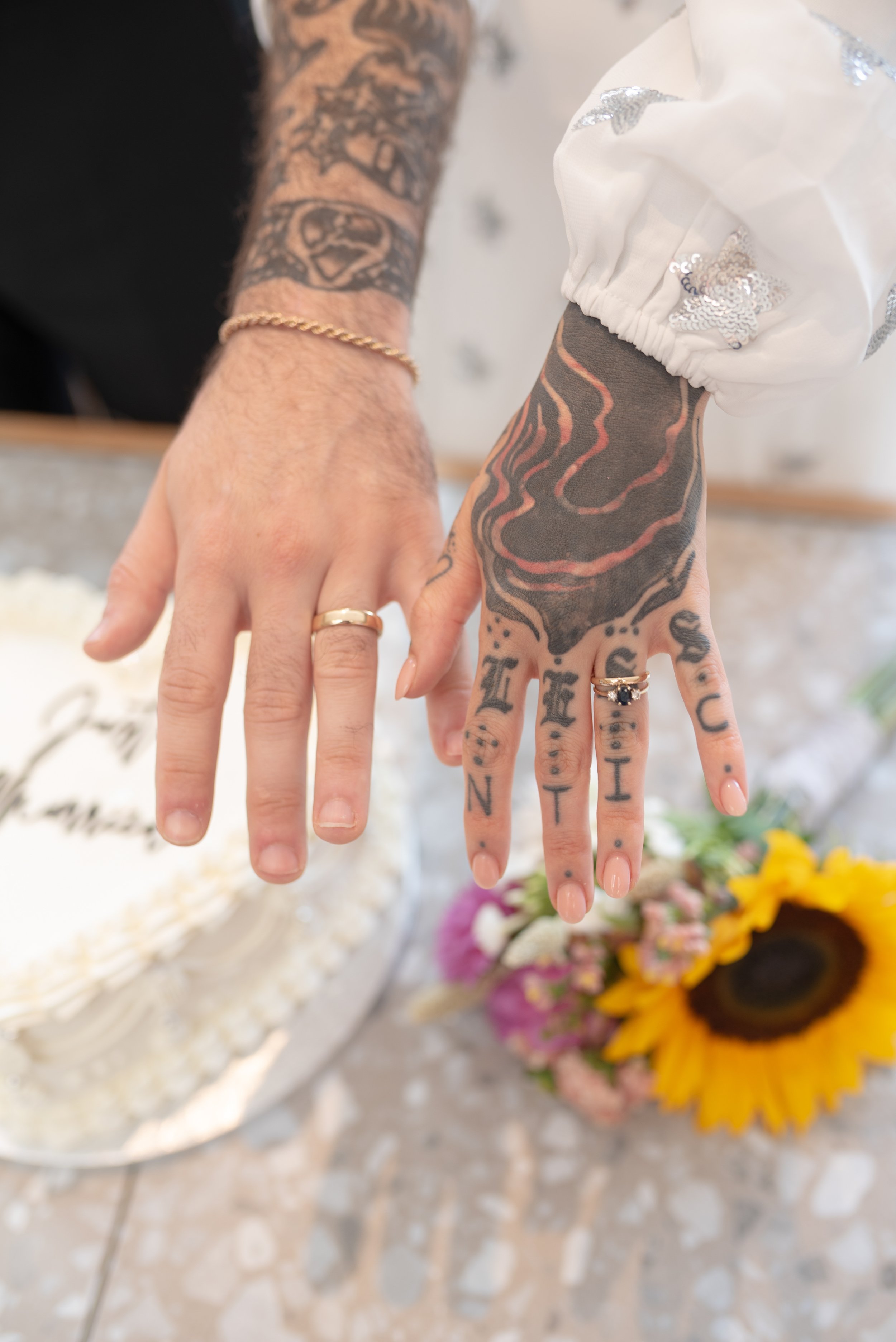 Close-up of a couple's hands showing wedding rings, with tattoos on their hands and arms, and a wedding cake and flowers in the background.