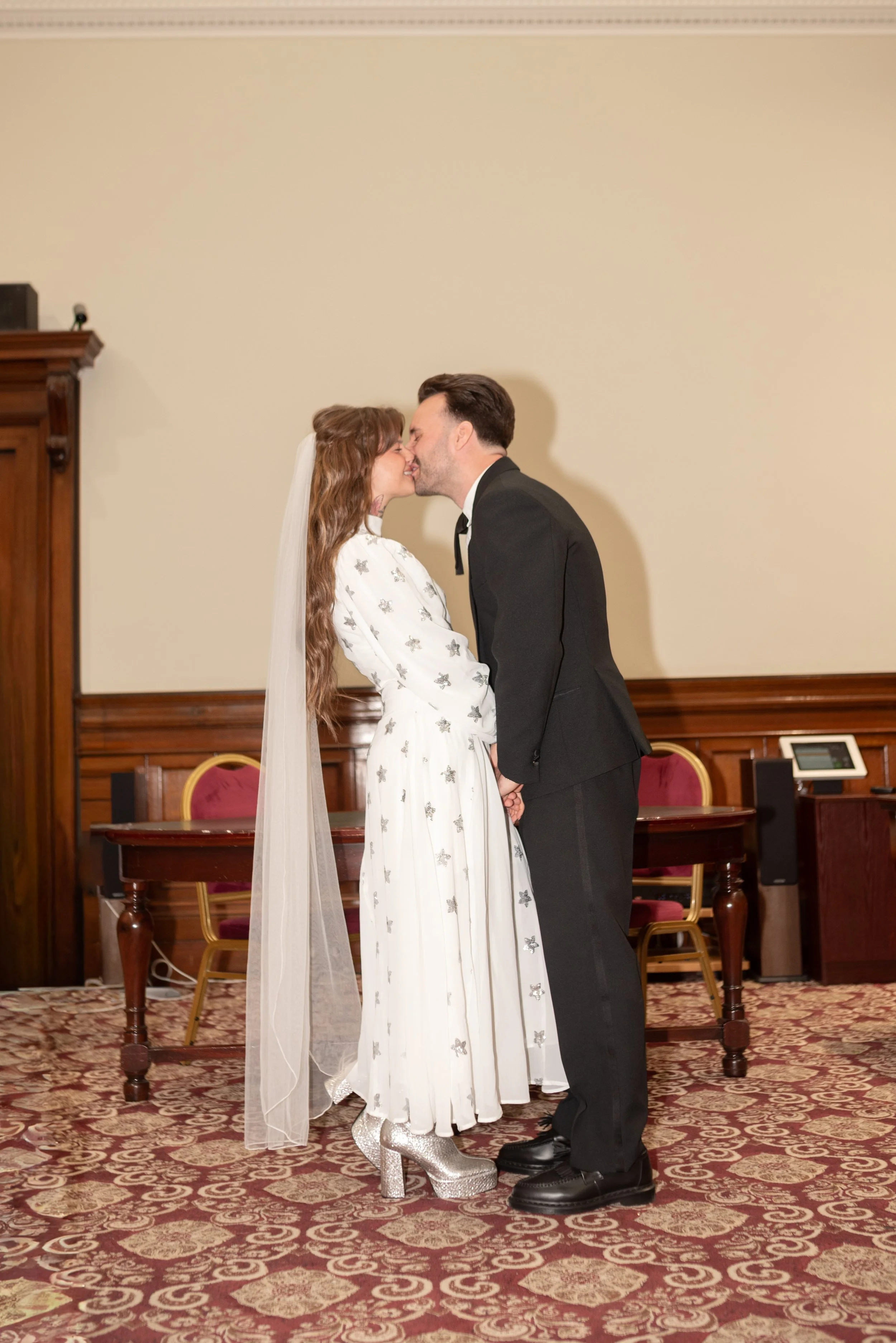 A bride and groom sharing a kiss during their wedding ceremony in a formal indoor setting.
