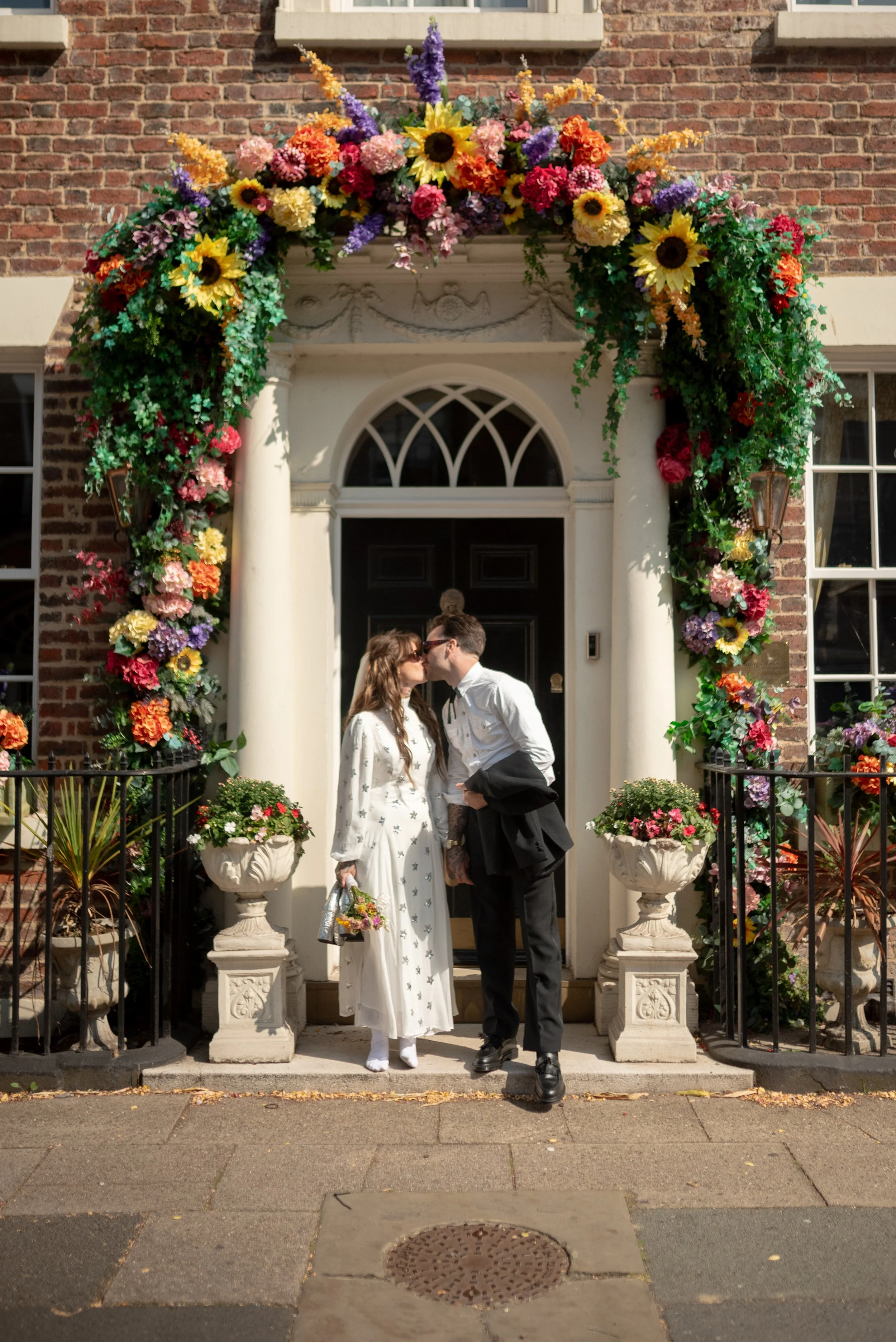 A couple sharing a kiss in front of a decorated doorway with a colorful floral arch and potted plants.
