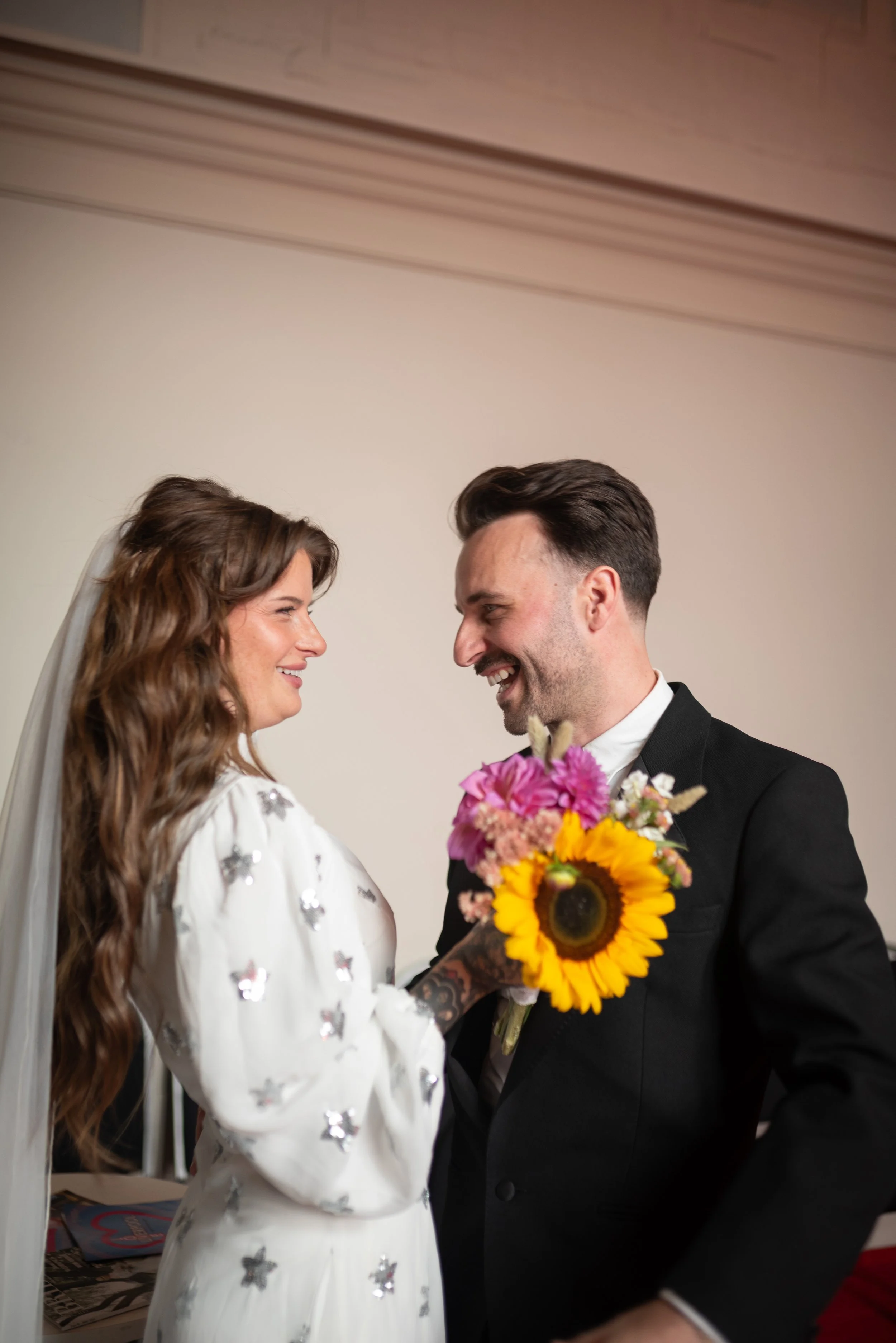 A bride and groom smiling at each other, with the bride holding a colorful bouquet of flowers, including a sunflower, in a room with a plain beige wall.