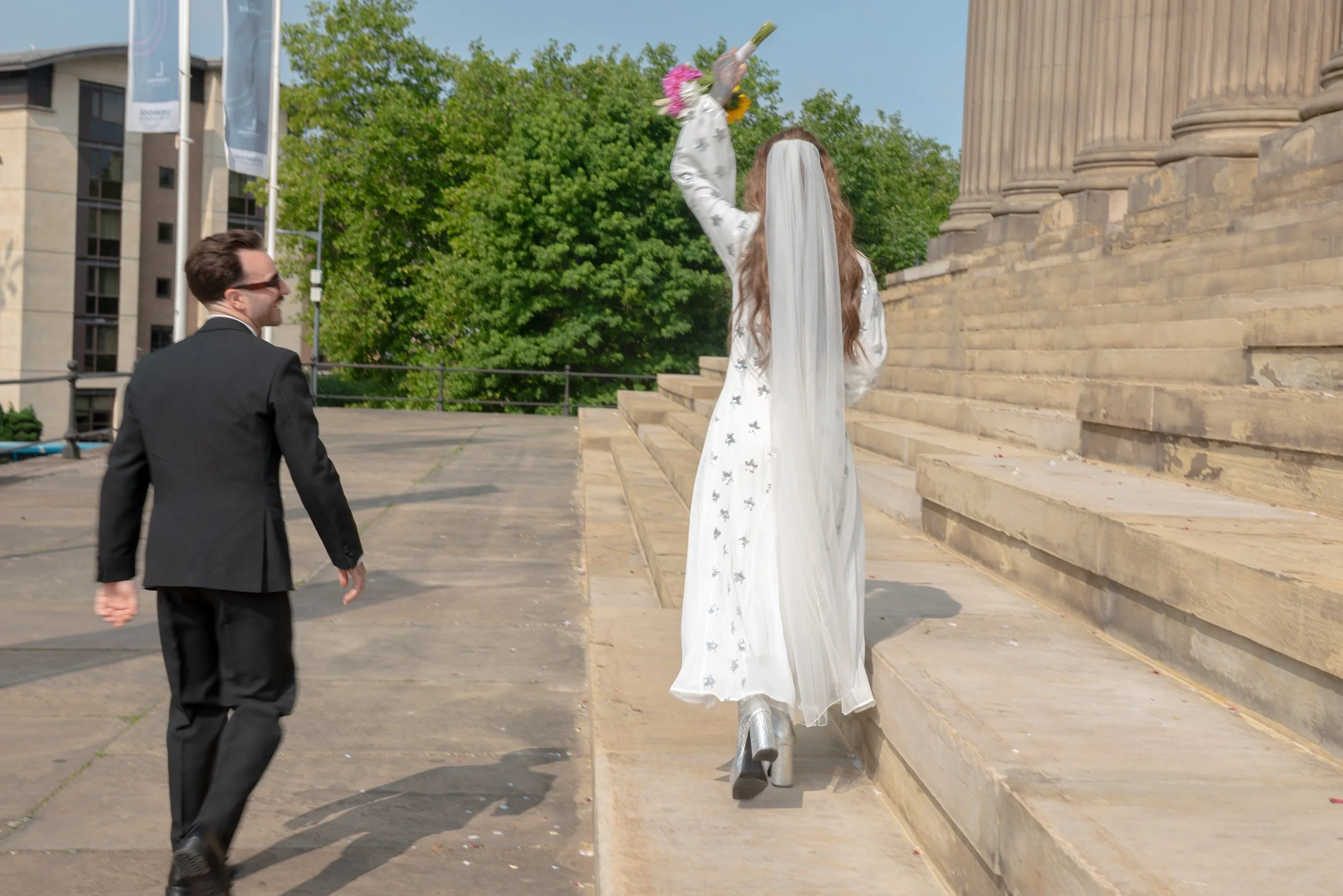 A woman dressed as a bride, wearing a white dress and long veil, is walking up the steps of a large historic building with columns, holding a bouquet. A man in a black suit and sunglasses is following her in an outdoor setting with trees and modern b